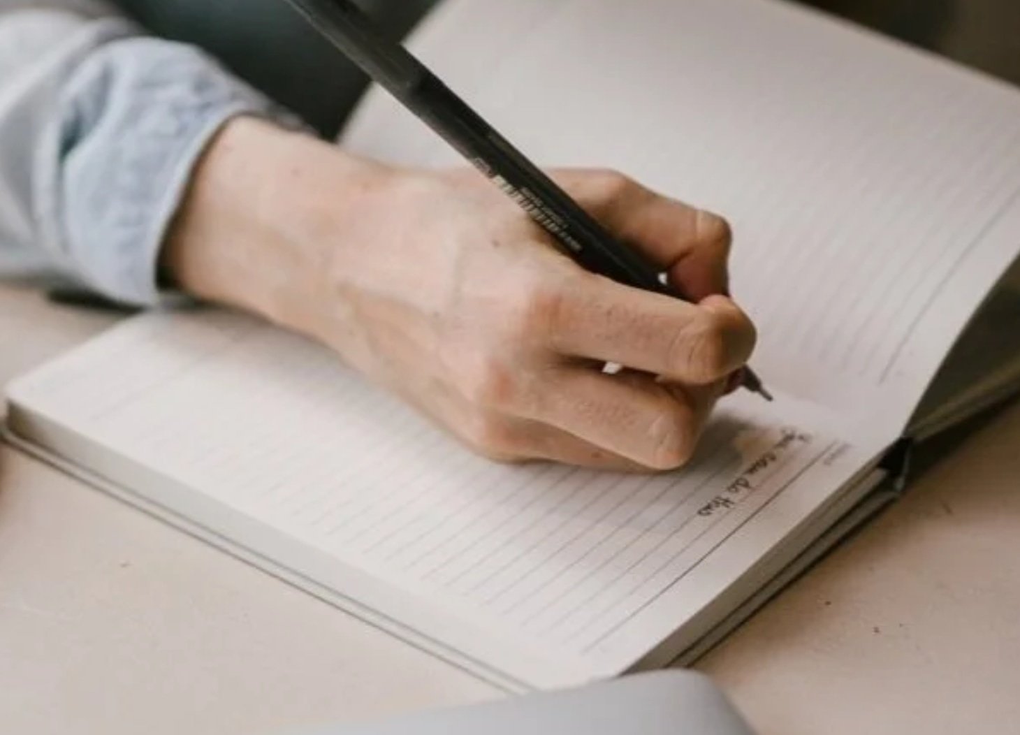 A person writes in a lined notebook with a black pen, only their hand and part of their arm visible.
