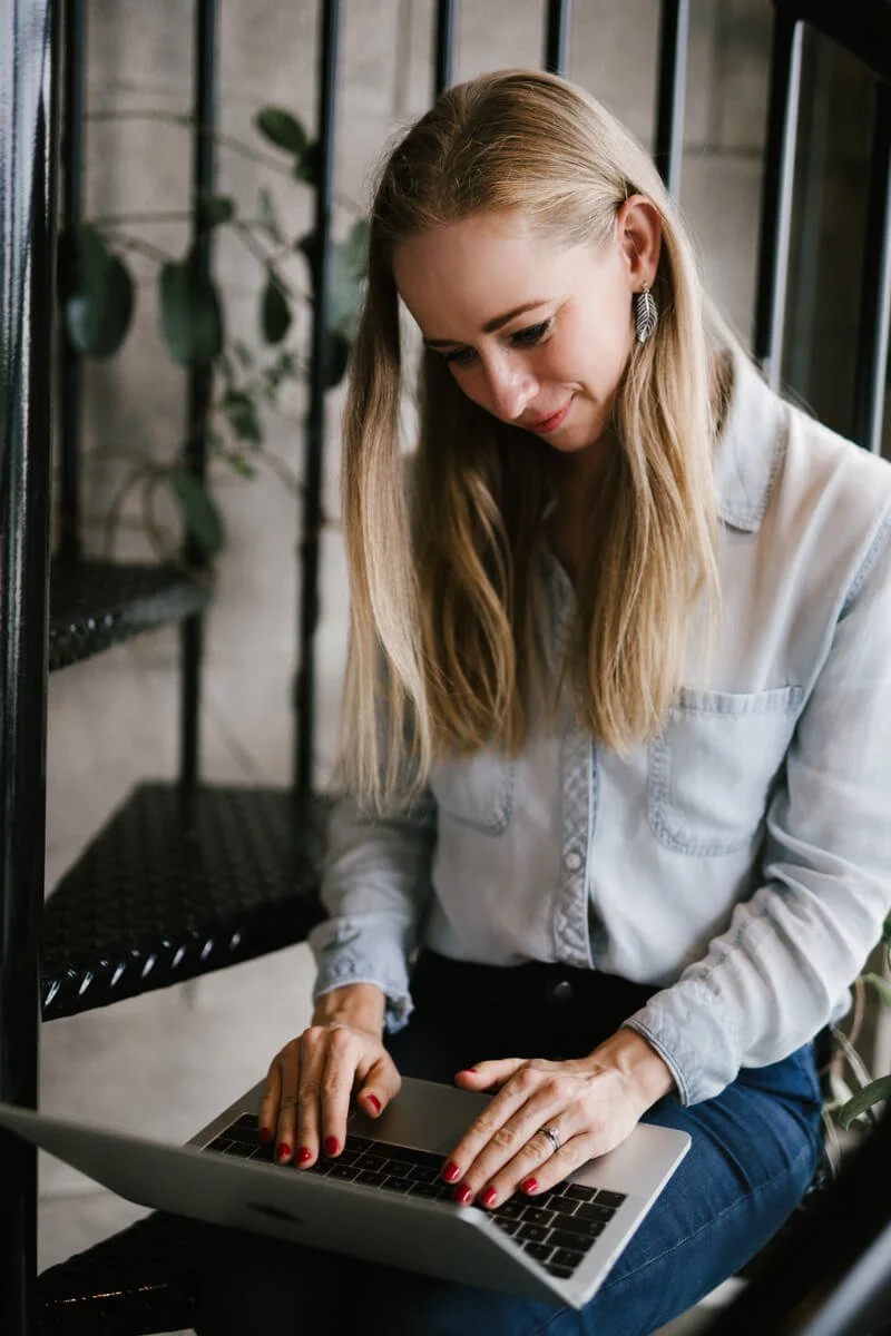 A woman with long blonde hair working on a laptop on her web design business while sitting near a window with black metal bars and a black shelf, in a cozy indoor setting.