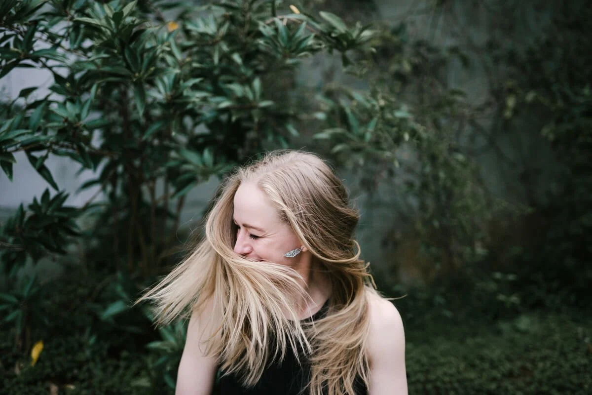 A young woman with long blonde hair is laughing or smiling while facing to the left, with her hair blowing across her face, standing outdoors in front of green foliage.