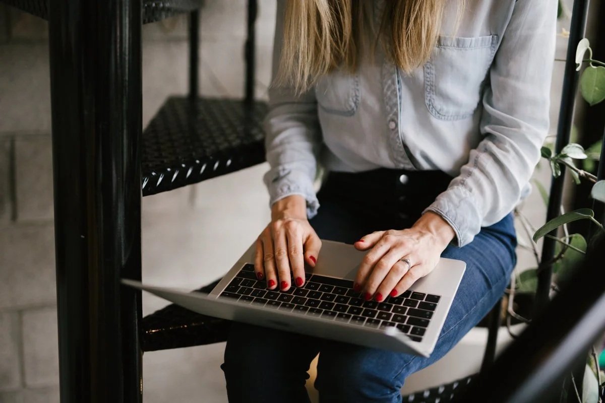 A woman sitting on a black metal chair with a laptop on her lap, wearing a light gray shirt and dark jeans, with red nail polish and a ring on her finger. She is working on her laptop.