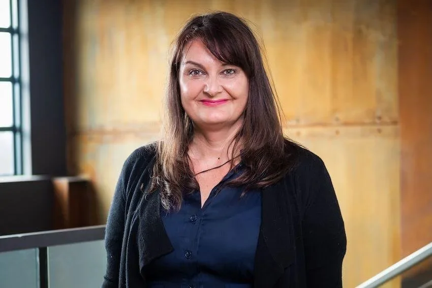 A woman with long brown hair and a black cardigan smiling in an indoor setting with warm wooden walls and a window.