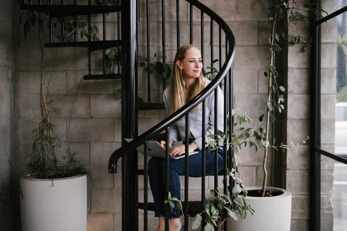 An Auckland Squarespace website designer sits on a spiral staircase with a laptop, surrounded by potted plants, gazing thoughtfully out a window.