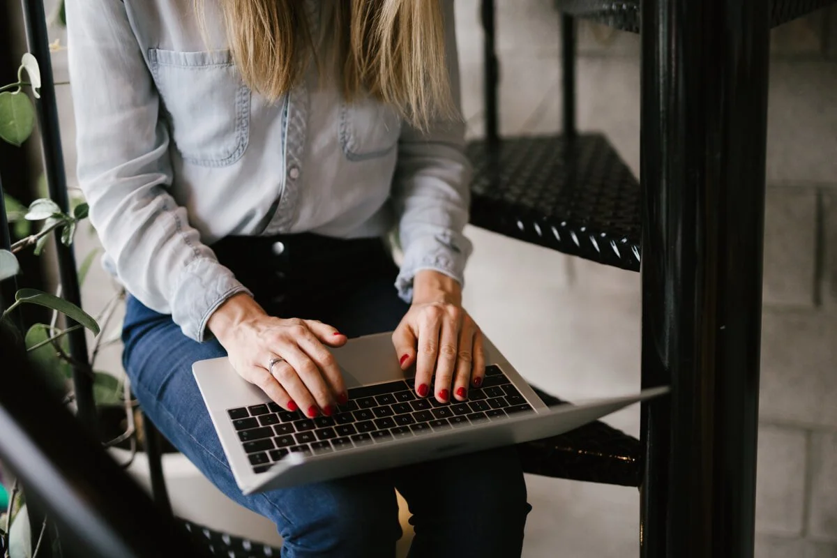 A web designer for wellness brands with long blonde hair, wearing a denim shirt and dark jeans, sitting on black metal stairs with a laptop on her lap, creating a health coach website design in Squarespace