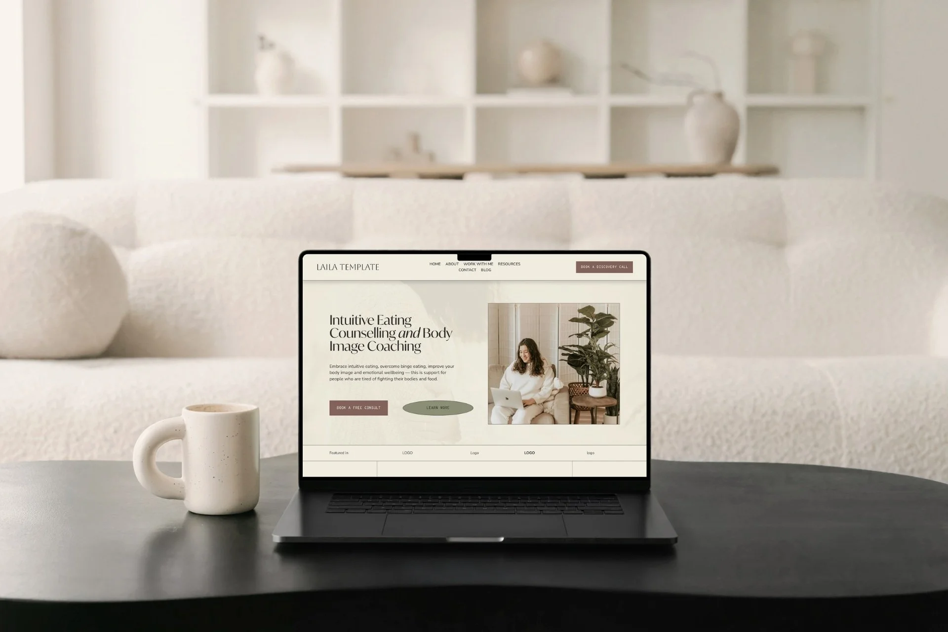 A laptop on a black table displays a wellness website by a skilled Squarespace website designer and copy editor; coffee mug, beige sofa, and shelves complete the scene.
