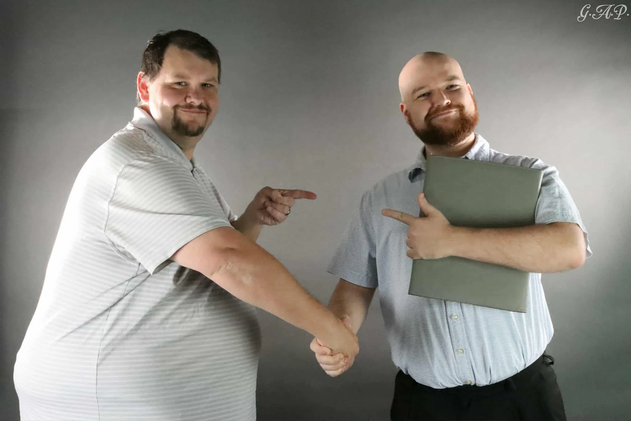 Two men shaking hands and pointing at each other, one is holding a laptop, both are smiling, standing against a plain gray background.
