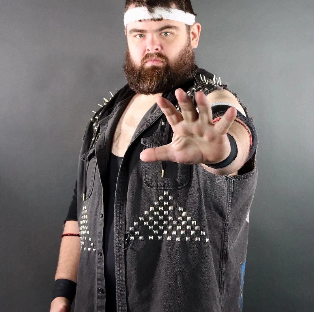 Man with a beard and wearing a white headband, sleeveless black denim vest with metal spikes, reaching outward with his hand, against a gray background.