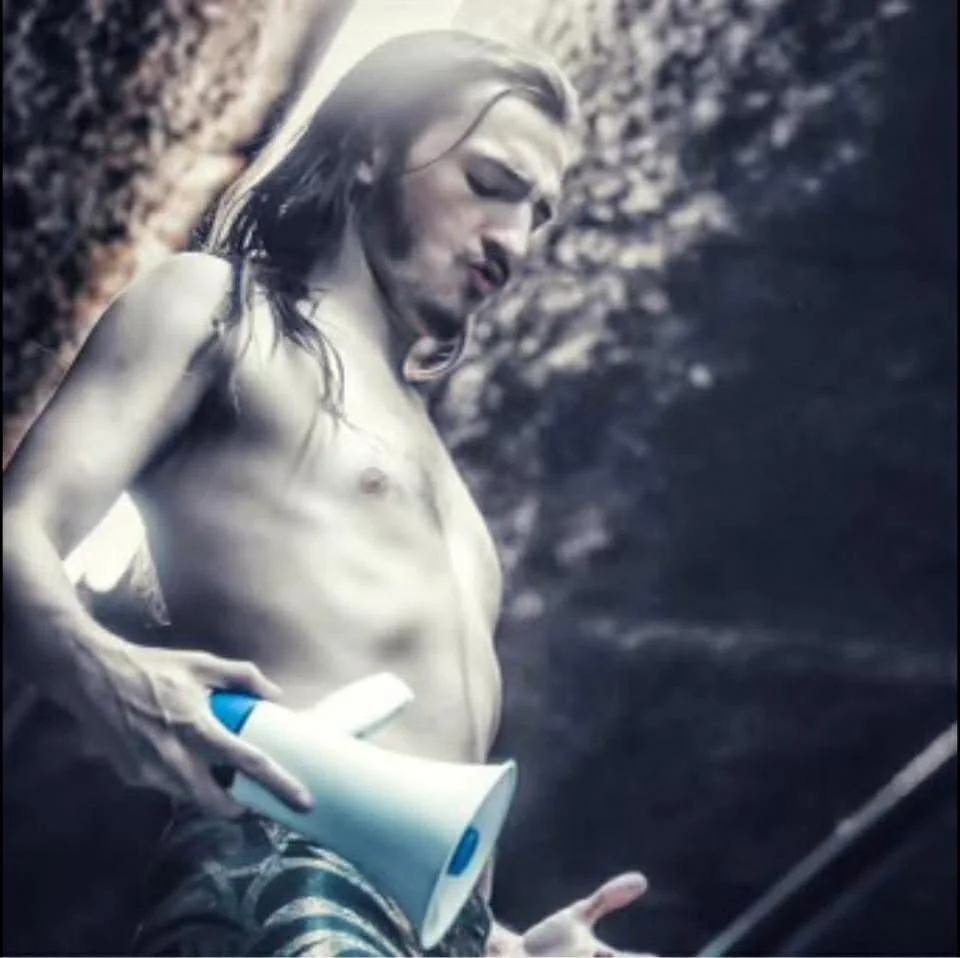 A shirtless old man with long hair standing outdoors near a rocky surface, holding a rolled-up paper cup and a long object, possibly a stick or cane.
