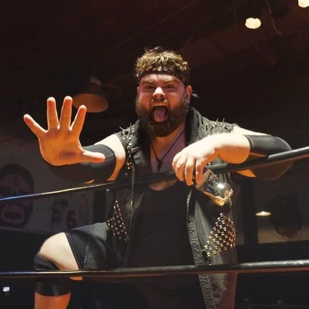 A man with a beard and curly hair wearing a spiked leather vest, making a playful face with his tongue out in a dimly lit venue.