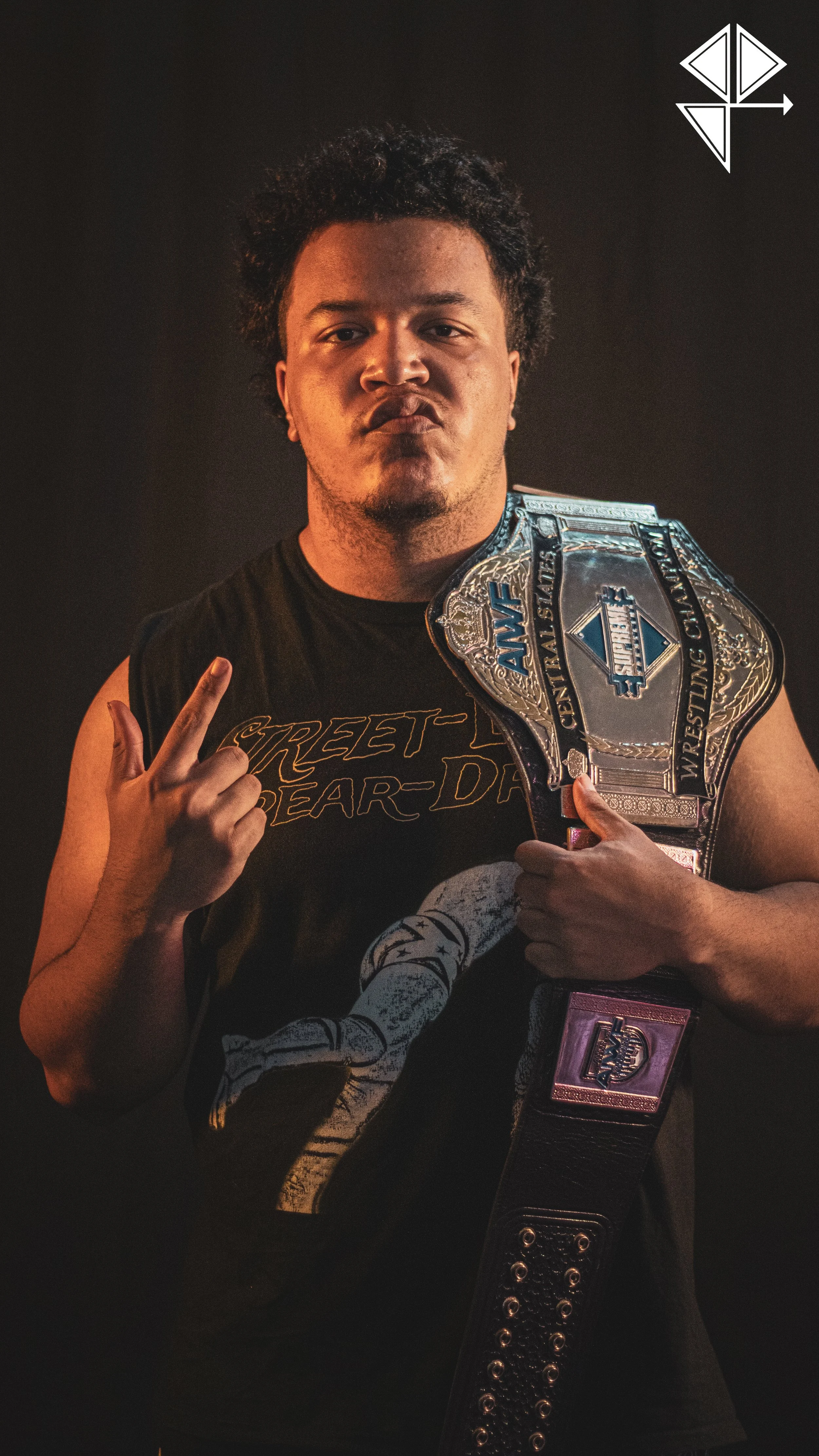 A male wrestler holding a championship belt over his shoulder, making a peace sign with his right hand and looking at the camera with a confident expression, against a plain dark background.