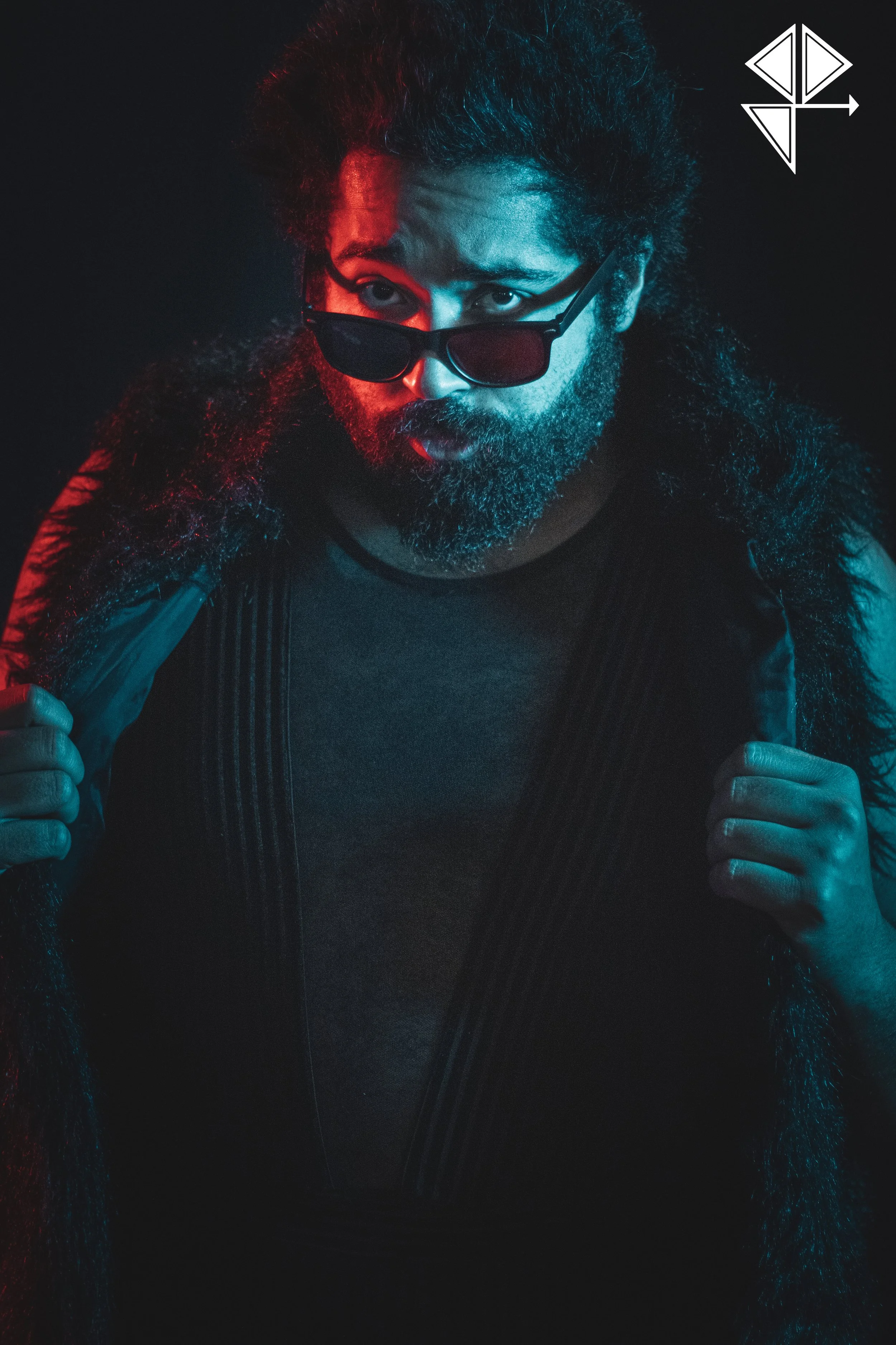 Man with glasses, sunglasses, long curly hair, and beard, wearing a dark shirt and vest, holding a fur-lined coat, with colorful lighting and a logo in the top right corner.