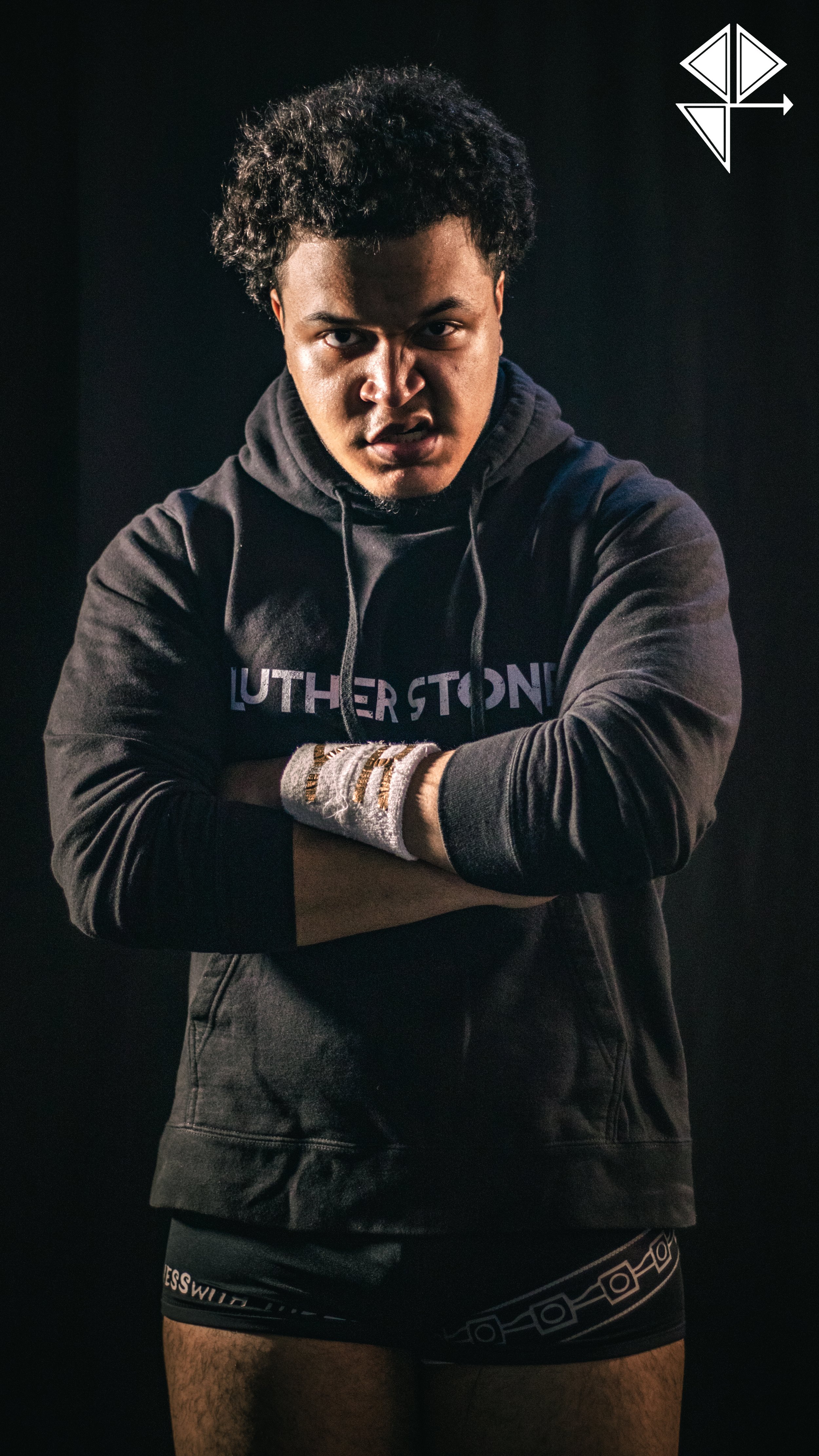 A young man with curly hair and an intense expression is posing against a dark background. He is wearing a black hoodie with the word 'LUTHERSTOWN' on it, gray and white fingerless gloves, and black underwear with a geometric pattern.
