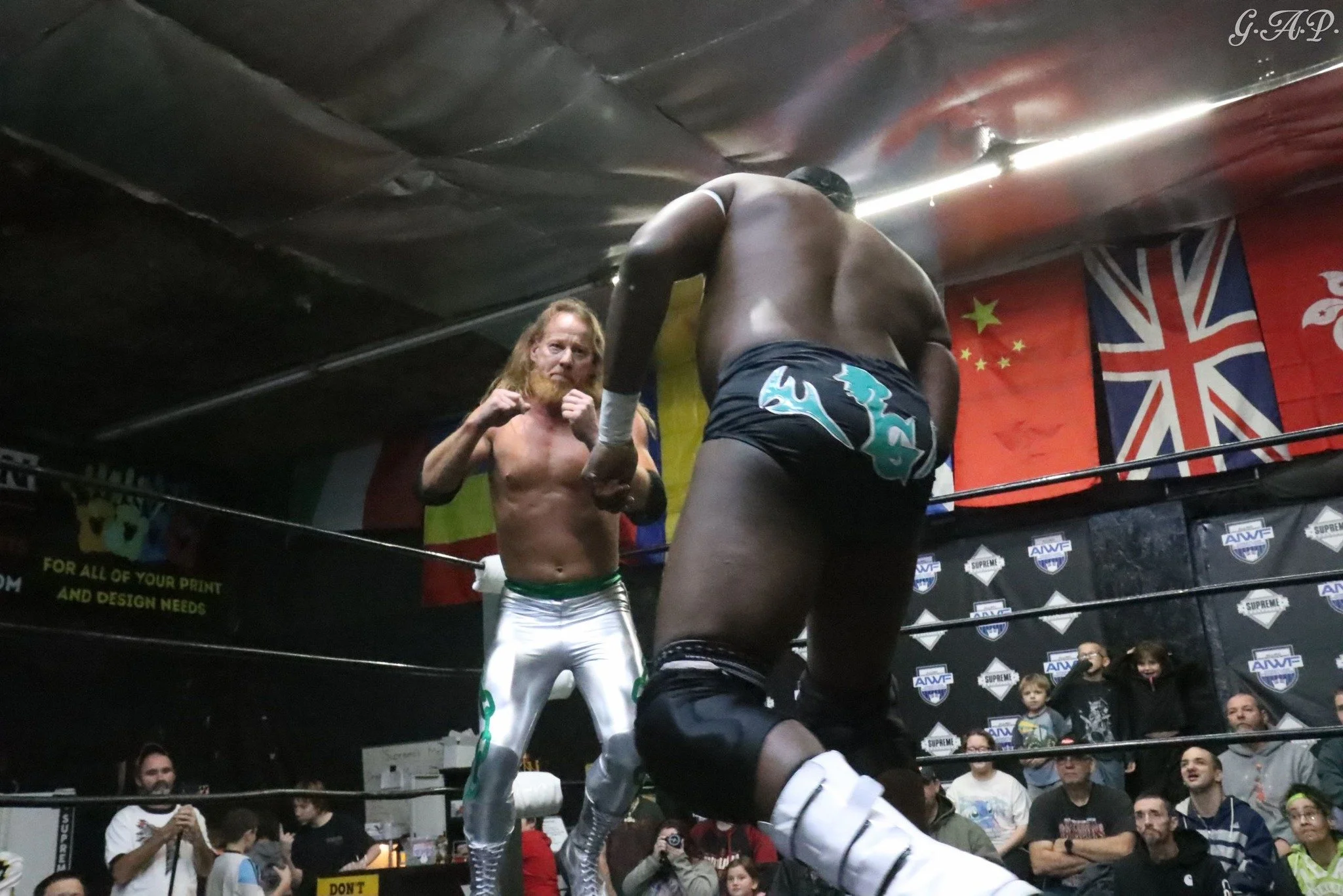 Two male wrestlers in a ring with an audience watching. The wrestler on the left has long red hair, a beard, and is wearing shiny white and green wrestling trunks. The wrestler on the right is larger, with dark skin, short hair, and is wearing black shorts with blue and white designs. Flags of China, the UK, and another country are hanging on the wall in the background.