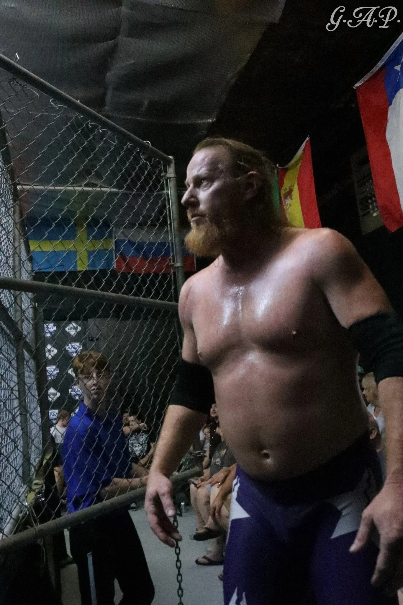 A shirtless wrestler with a beard and long hair in a wrestling arena, standing near a chain-link fence, with spectators in the background and flags hanging behind.