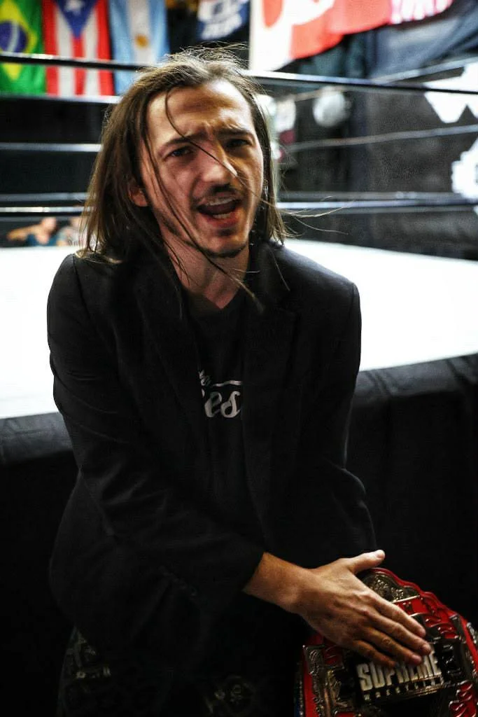 A man with shoulder-length brown hair, wearing a black jacket, is sitting in a wrestling ring with a waist holster or a plaque in his hand.
