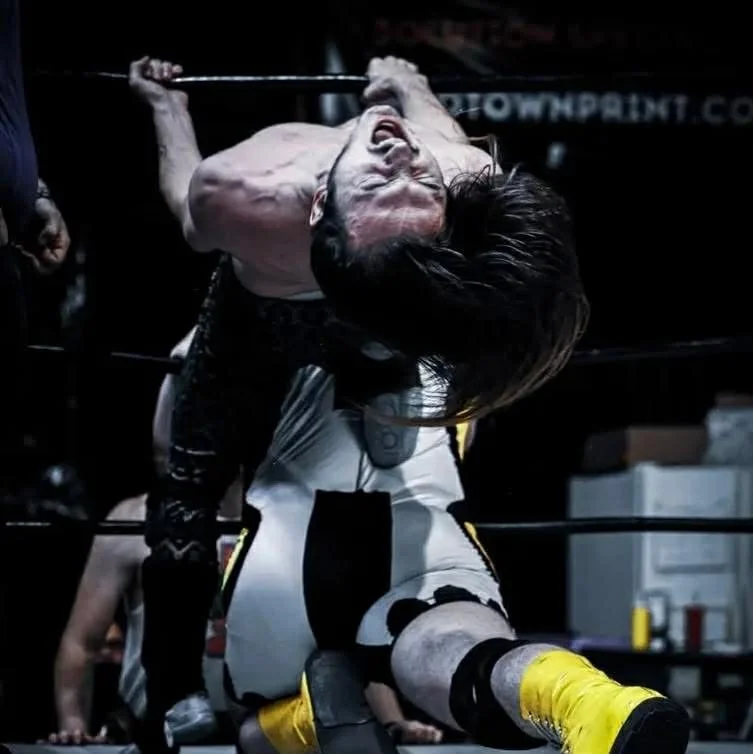 Person in wrestling ring, leaning backward with hair over face, gripping the top rope with one hand, in an intense wrestling move.