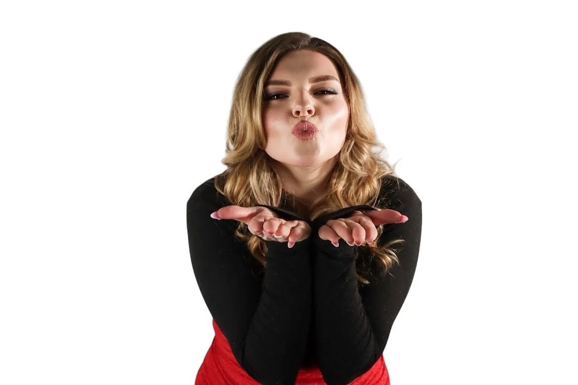 Woman with wavy blonde hair blowing a kiss toward the camera, wearing a black top and red skirt, against a white background.