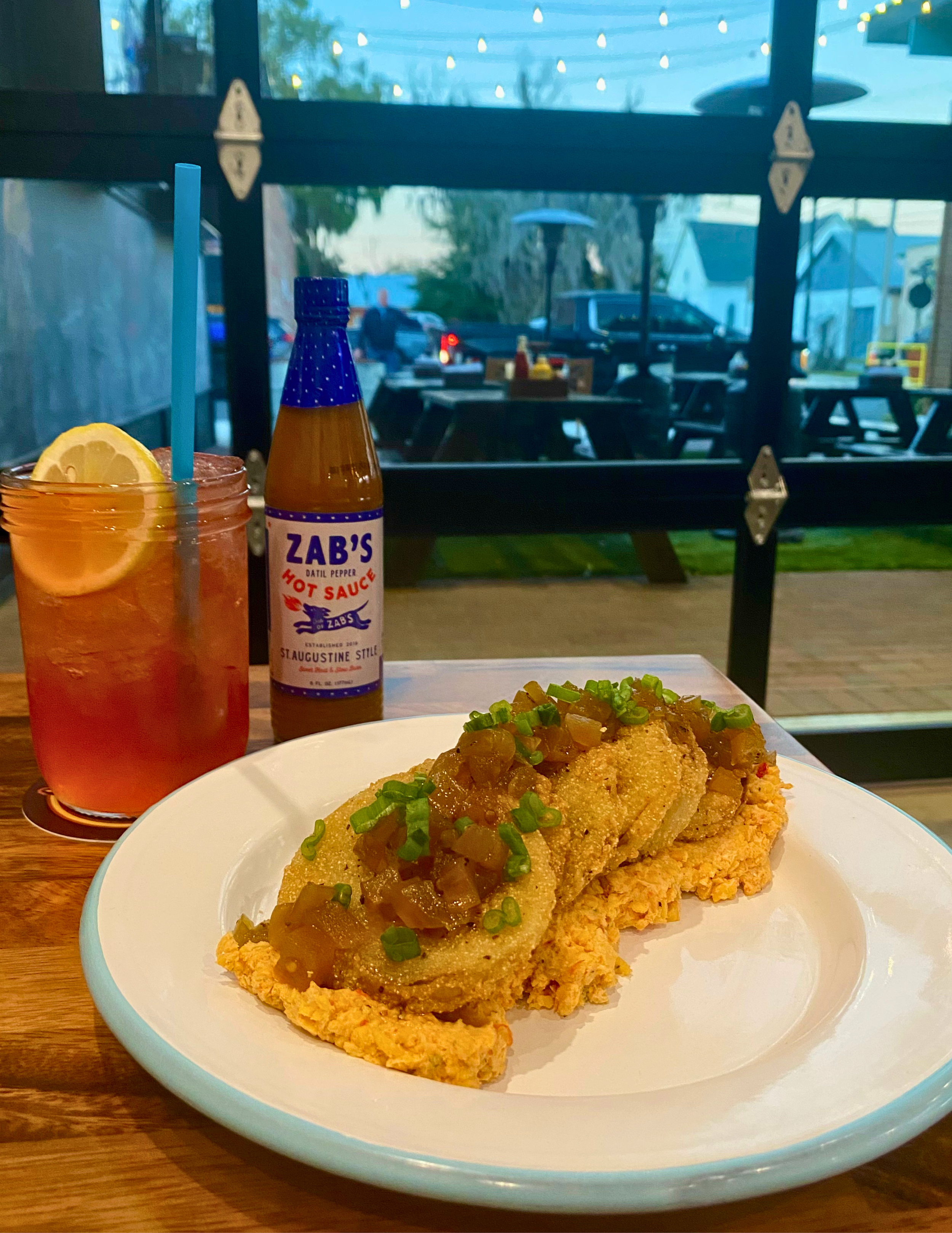 A plate of fried fish topped with green onions and sauce, with a glass of iced tea with lemon and a straw, and a bottle of Zabs Hot Sauce, on a wooden table in a restaurant with a large window showing outdoor seating and string lights.