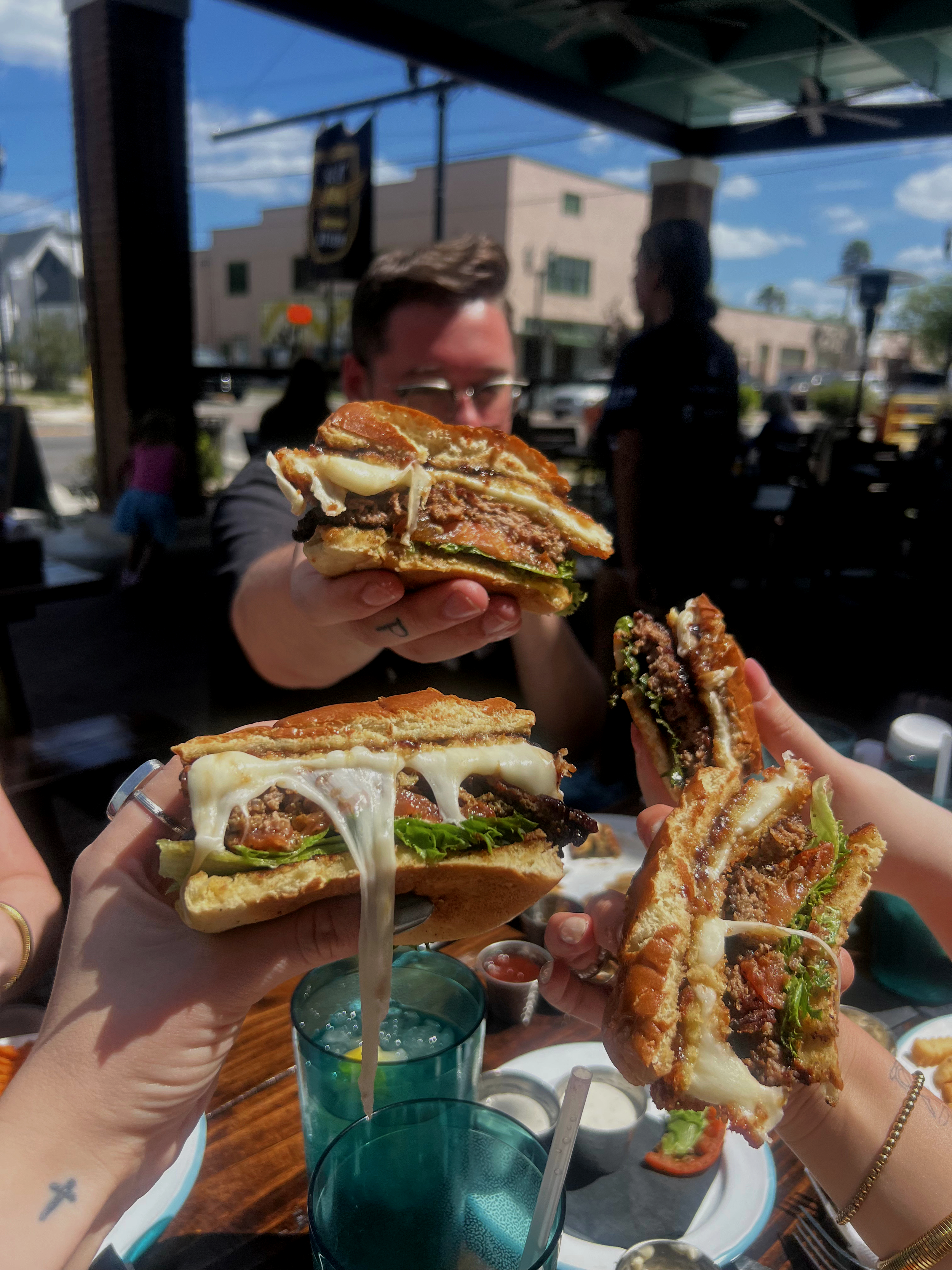 People holding up various slices of burger with melted cheese, vegetables, and meat at an outdoor restaurant on a sunny day.