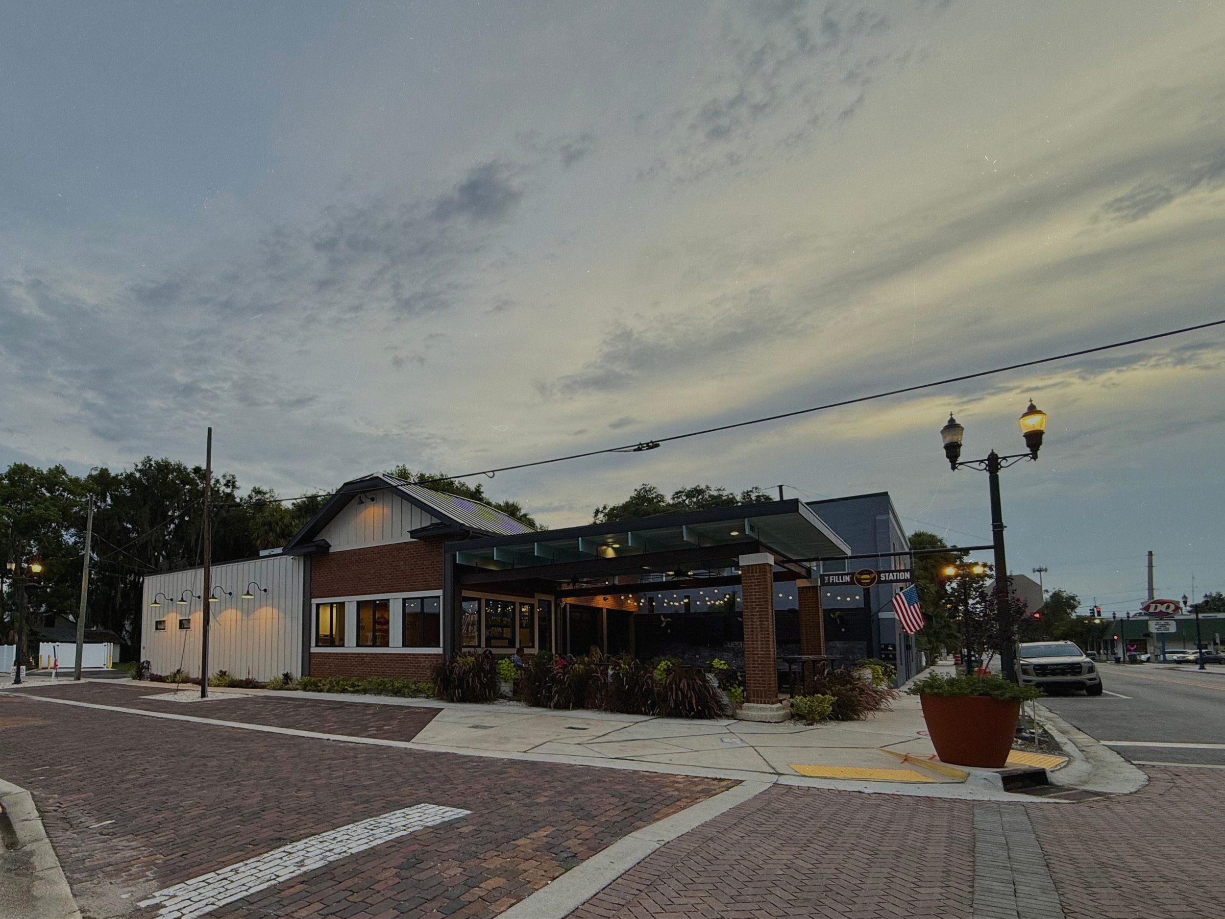 A white and brick building with a gabled roof, large windows, and a star emblem on the front, situated in a sunny area with a parking lot and a sidewalk, under a clear blue sky with some clouds.