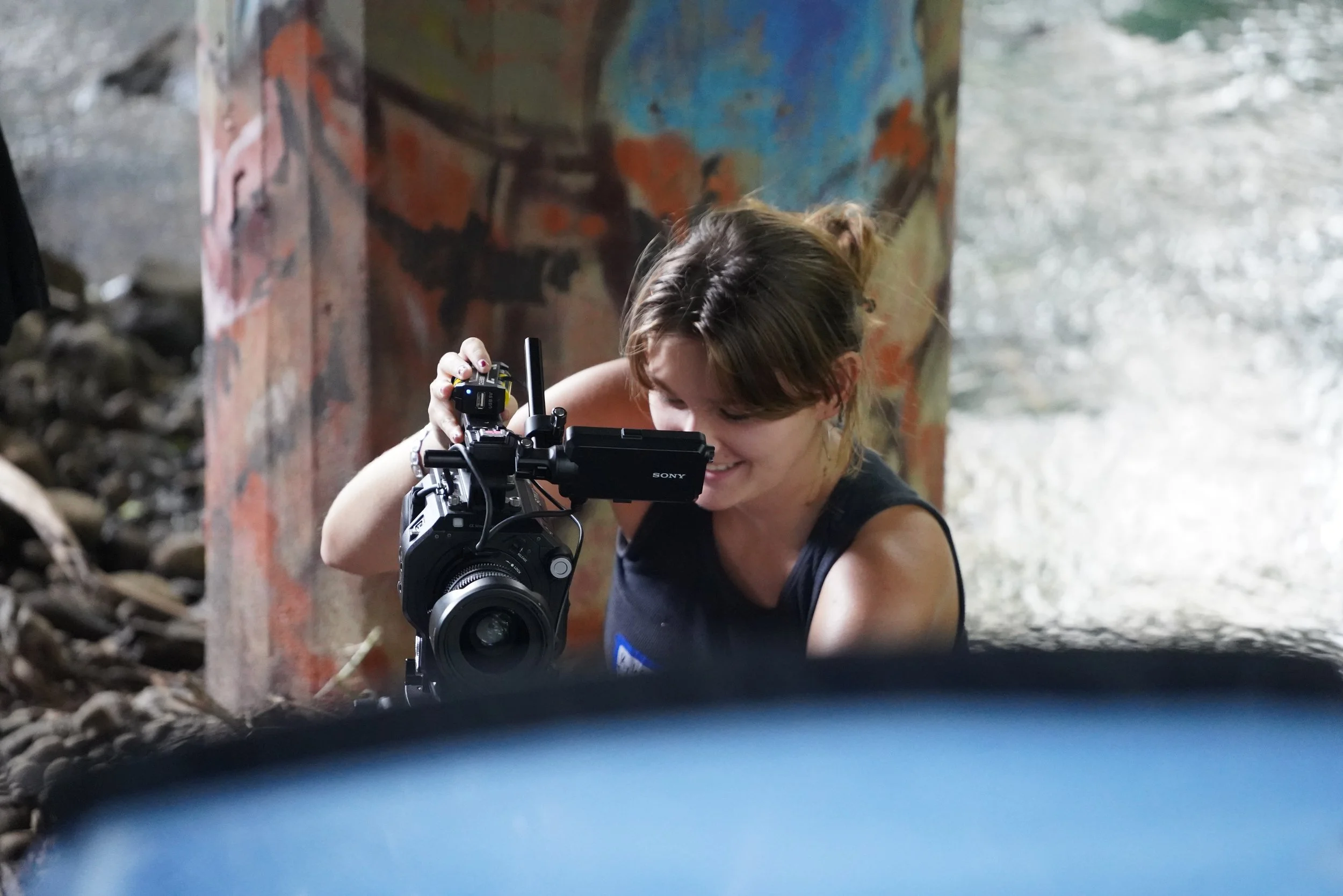 A woman is looking through the viewfinder of a professional video camera under a bridge or tunnel with graffiti on the wall of the structure.