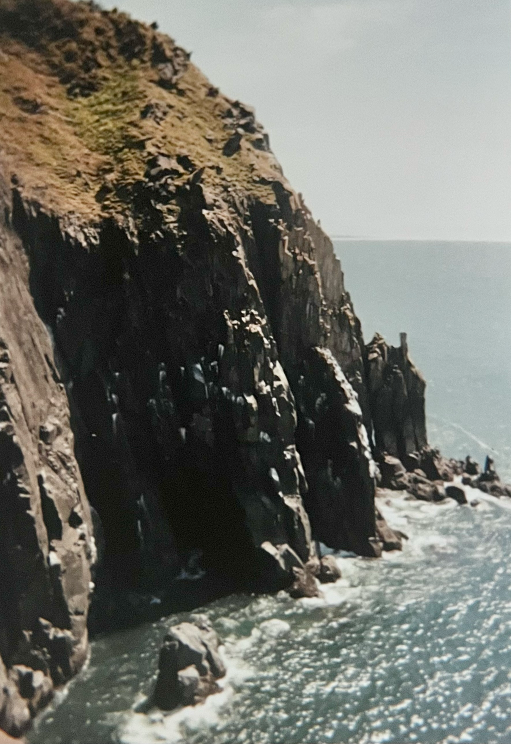 Cliffside view of rugged black rocks with ocean waves crashing at the base and a cloudy sky in the background.