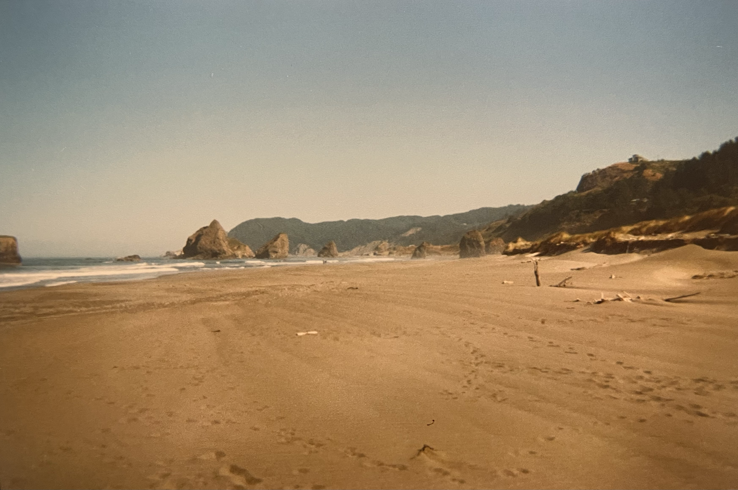 A wide sandy beach with footprints, rocks in the water, and distant hills under a clear sky.