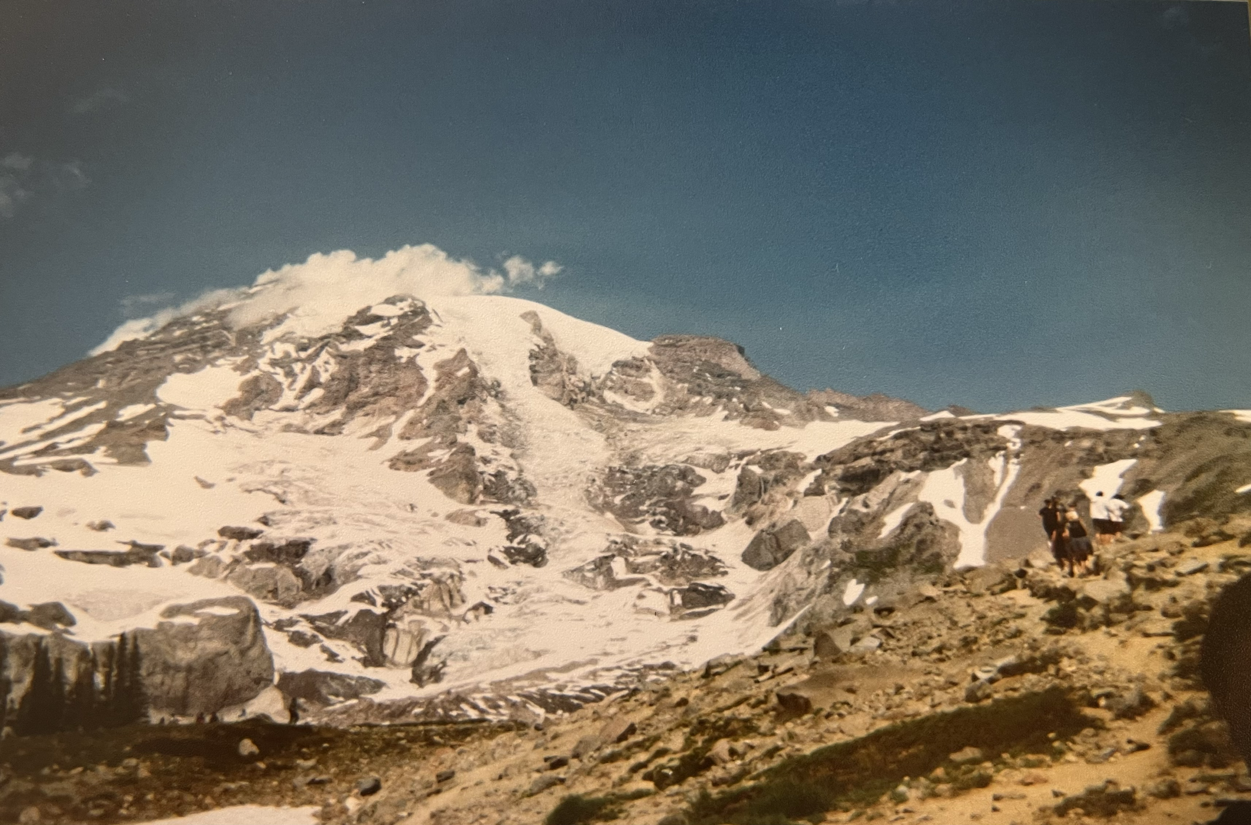 Snow-capped mountain with rocky terrain and a small group of hikers with backpacks walking along a trail.