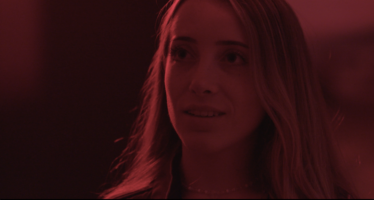 A woman with long wavy hair looking slightly to the side with a surprised or curious expression, illuminated by red lighting.