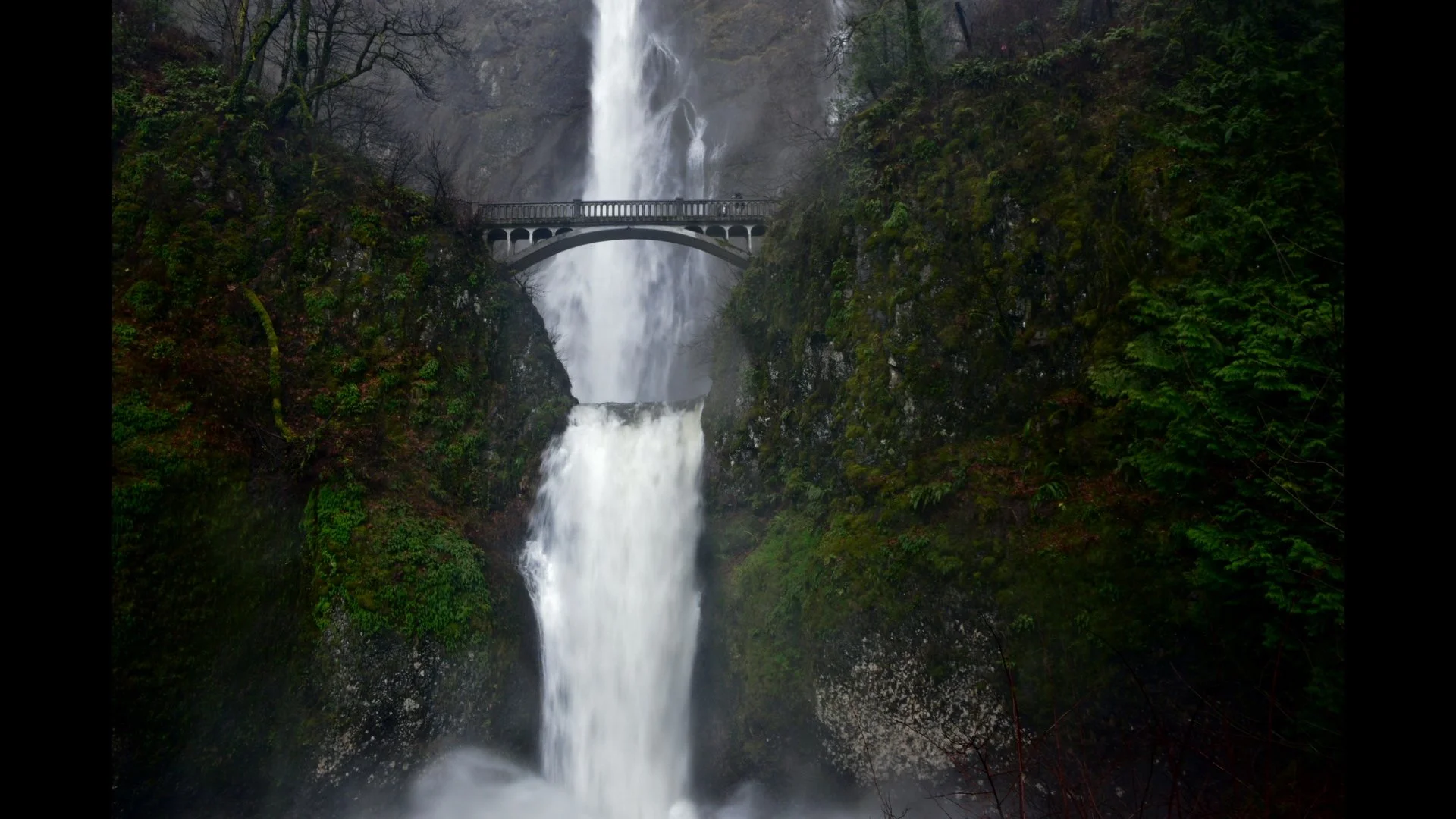 A tall waterfall flowing over rocks in a lush green forest, with a bridge over the waterfall near the top.