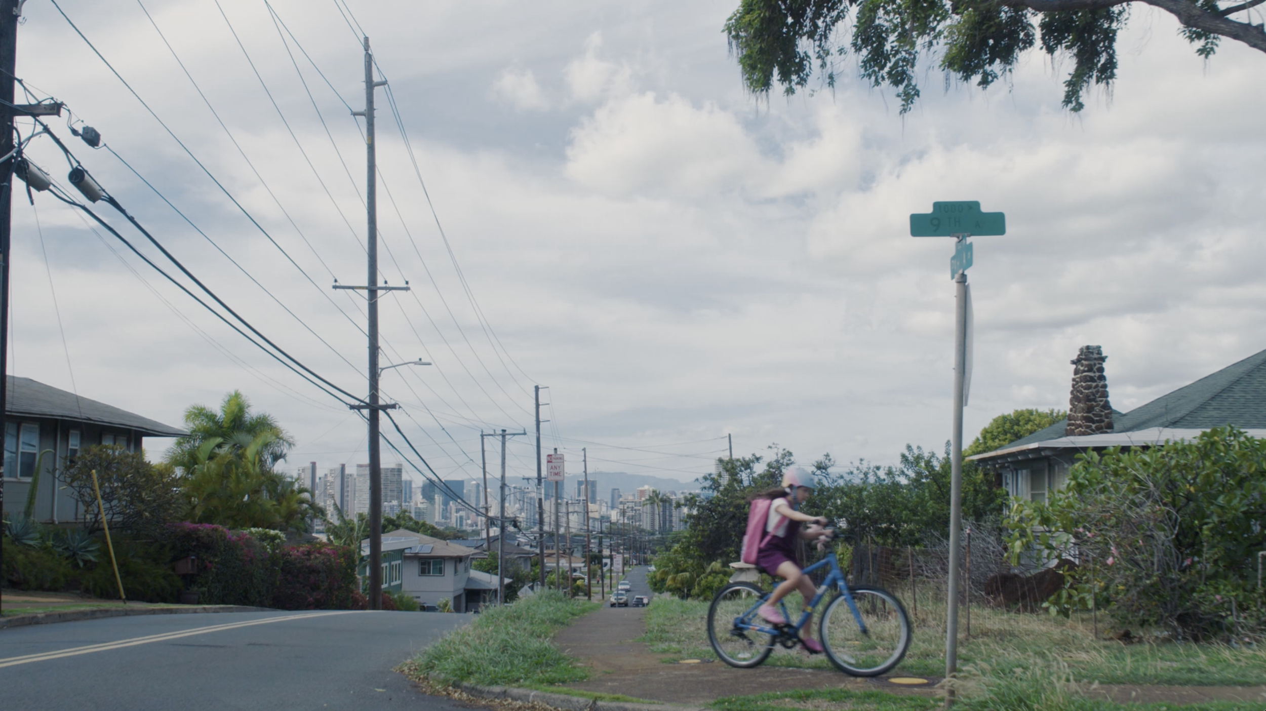 A young girl riding a bicycle on a neighborhood street with houses, power lines, and a city skyline in the background.