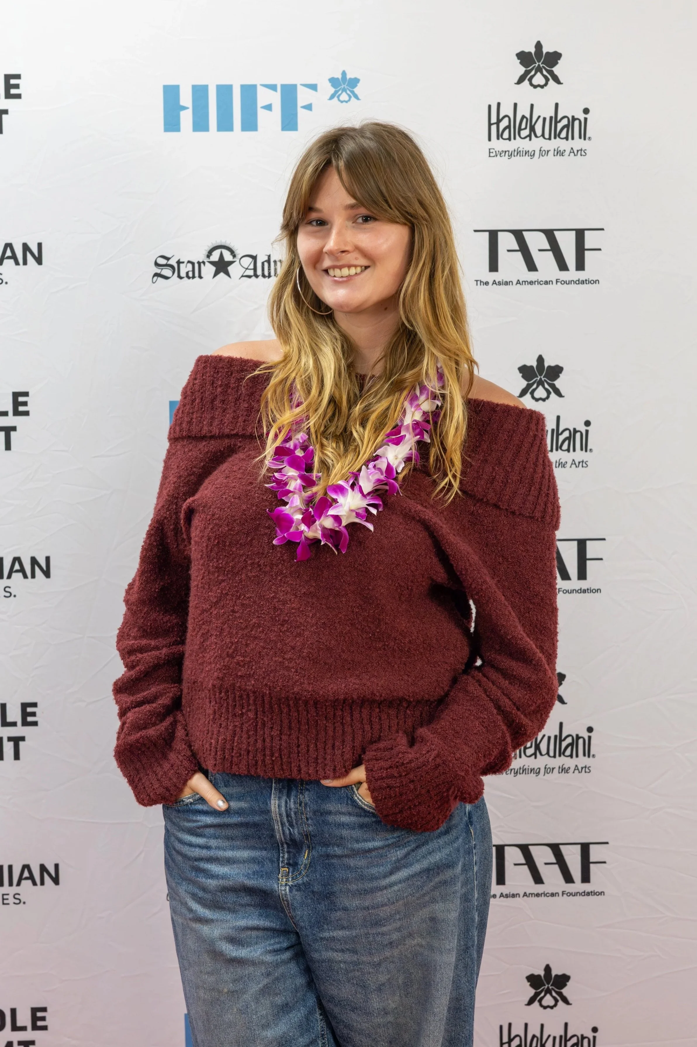 A young woman wearing a maroon off-shoulder sweater and blue jeans, smiling in front of a step and repeat backdrop with logos of the Asian American Foundation and other sponsors, and wearing a purple and white lei.