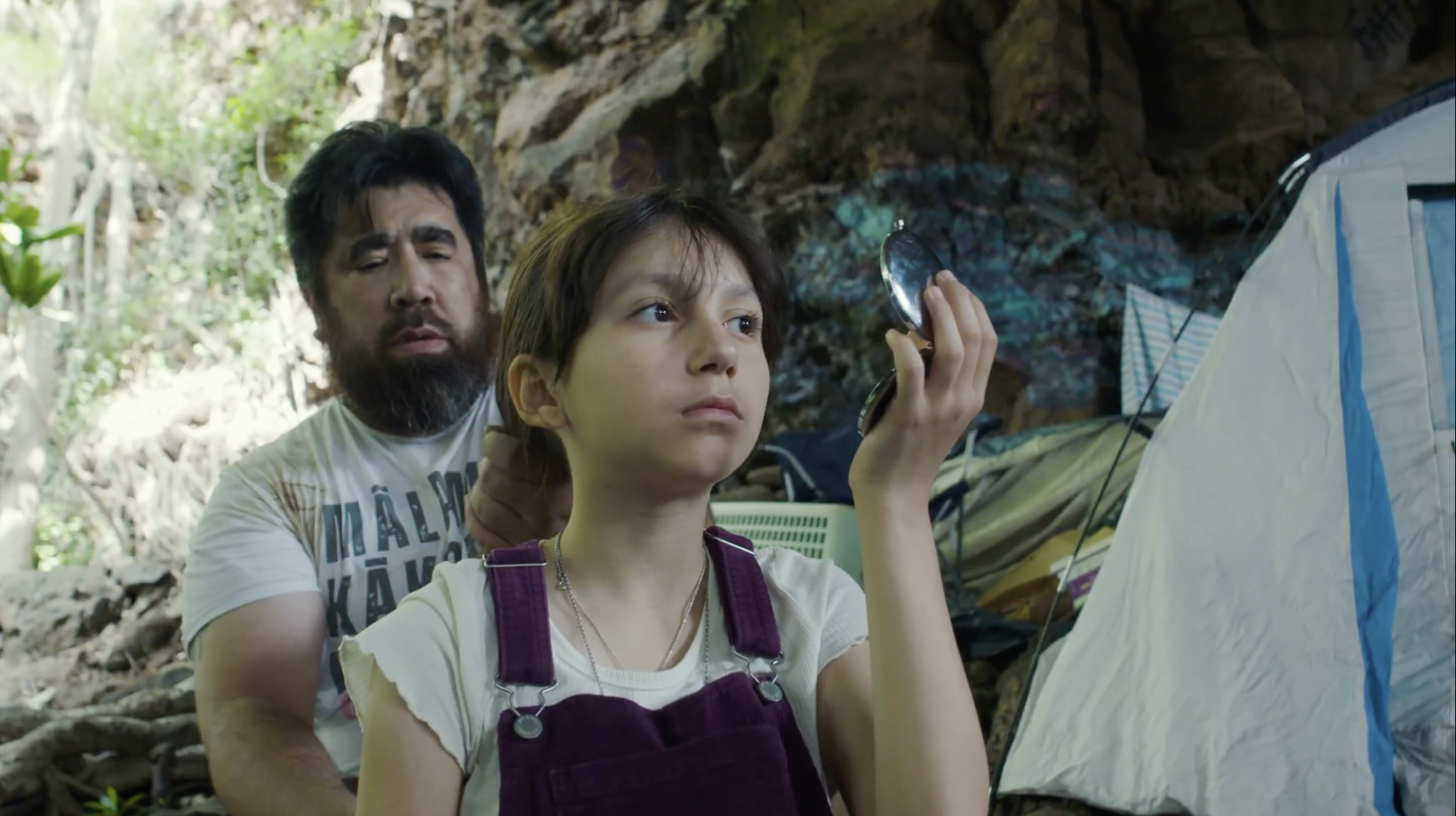 A young girl holding a small mirror in a forest, with a man standing behind her near a tent.