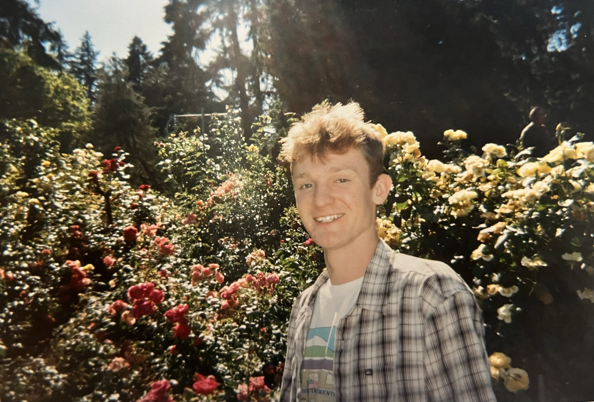 A young man with short, light brown hair and a checkered shirt smiling in a colorful garden filled with yellow, pink, and red flowers on a sunny day.