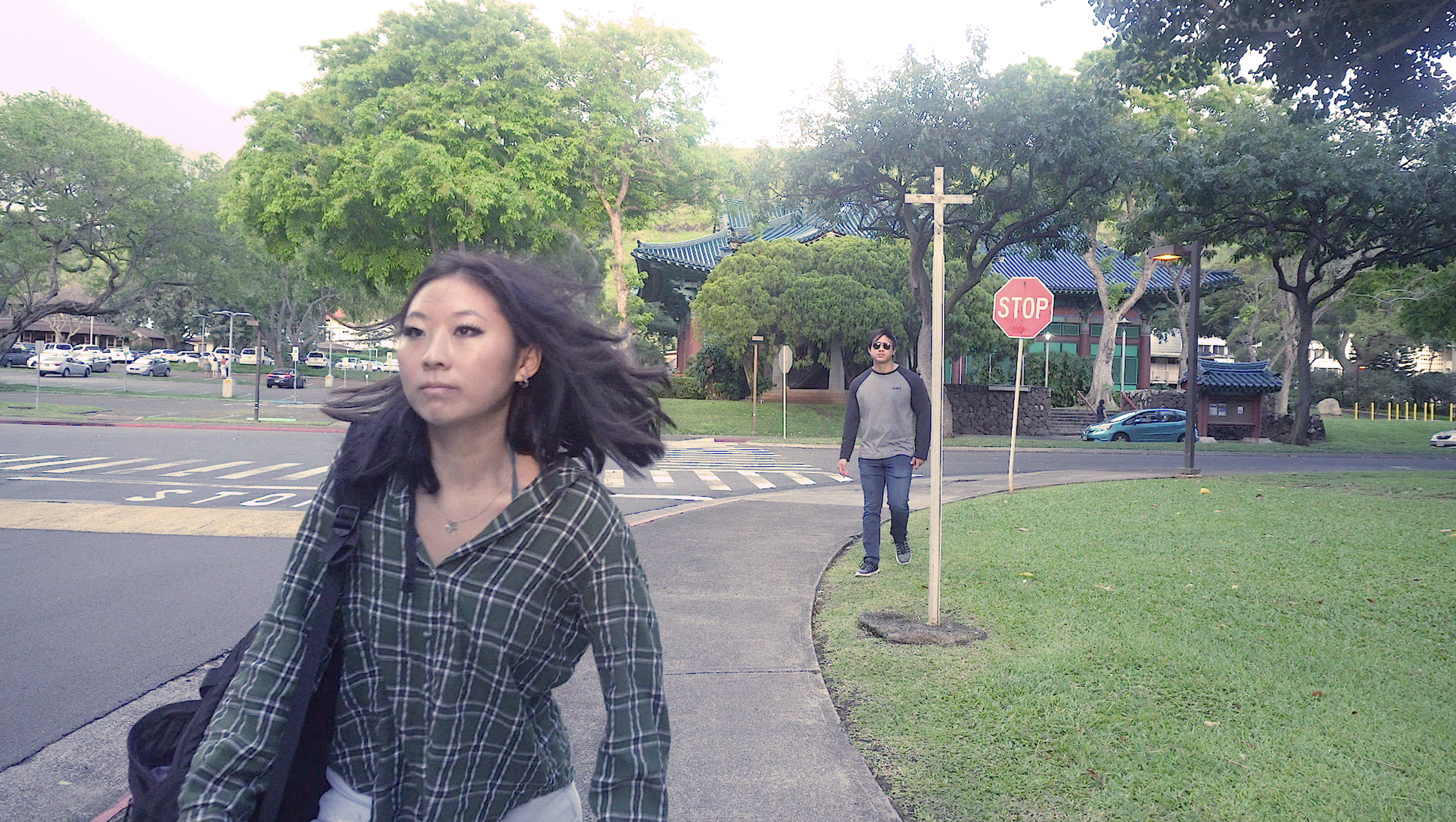 Two people walking on a sidewalk near a stop sign, with trees, parked cars, and a building with a traditional Asian-style roof in the background.