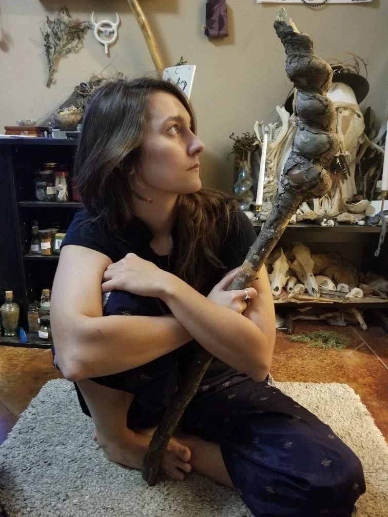 Woman sitting on a rug holding a large, twisted root or gnarled wooden stick in a room filled with skeletal remains and bottles.