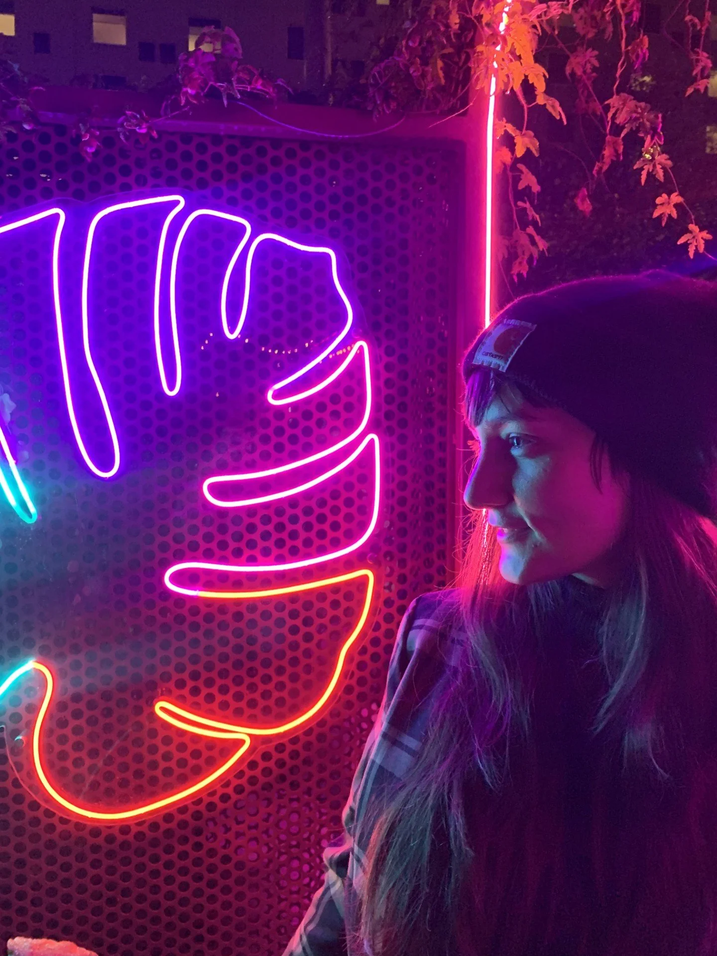 A woman with long hair wearing a beanie hat and checkered shirt looking at a bright neon sign of a tropical leaf in purple, pink, red, and orange colors.
