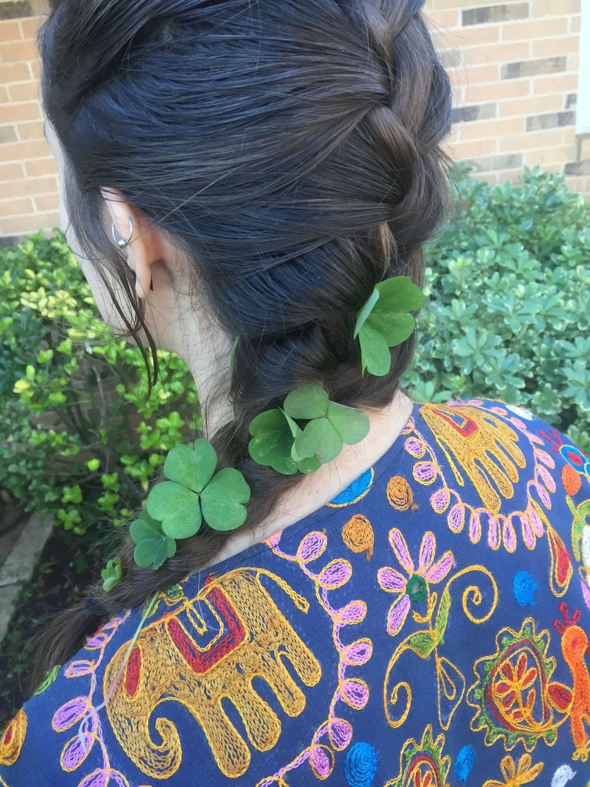 A woman with dark hair styled in a braid decorated with green clover leaves, wearing a colorful embroidered dress, standing outdoors near green plants and a brick wall.