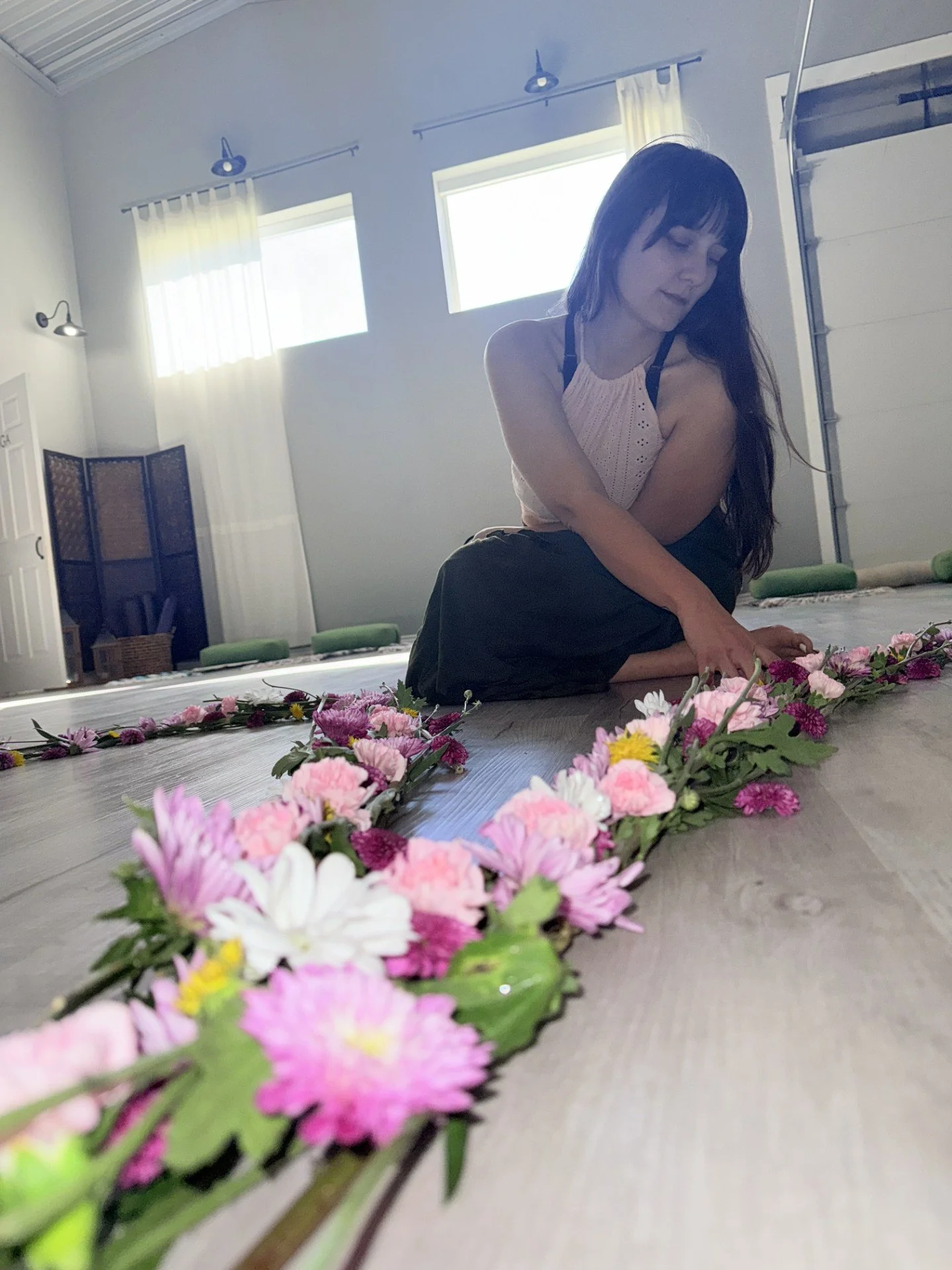 A woman sitting on the floor arranging pink and white flowers in a line on a wooden surface in a bright room with white walls and windows.