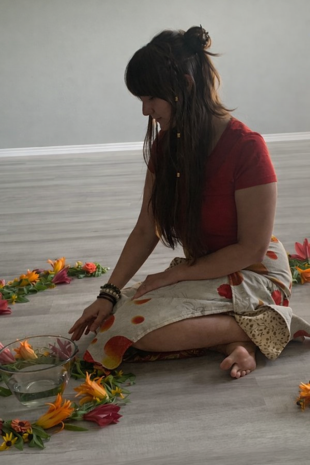 A woman with long, dark hair and wearing a red shirt and patterned skirt is sitting cross-legged on the floor surrounded by colorful flowers and a glass bowl of water. She is reaching towards the flowers with her left hand.