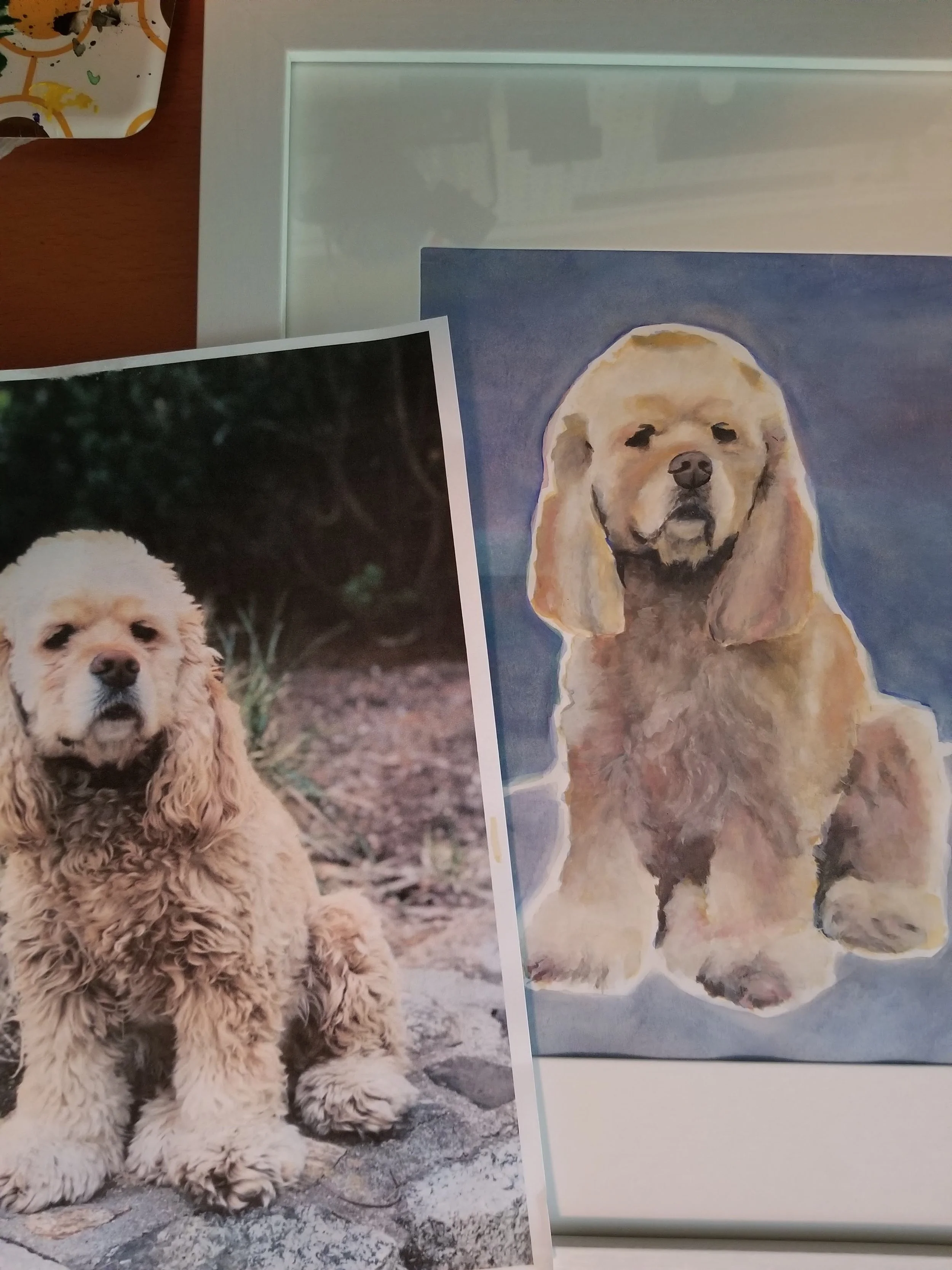 Photograph of a realistic painting of a golden-colored Cocker Spaniel puppy sitting outdoors, with a physical print of the same puppy placed in front of the painting.
