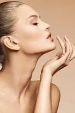 A woman with blonde hair and makeup posing with her hand near her face against a plain background.