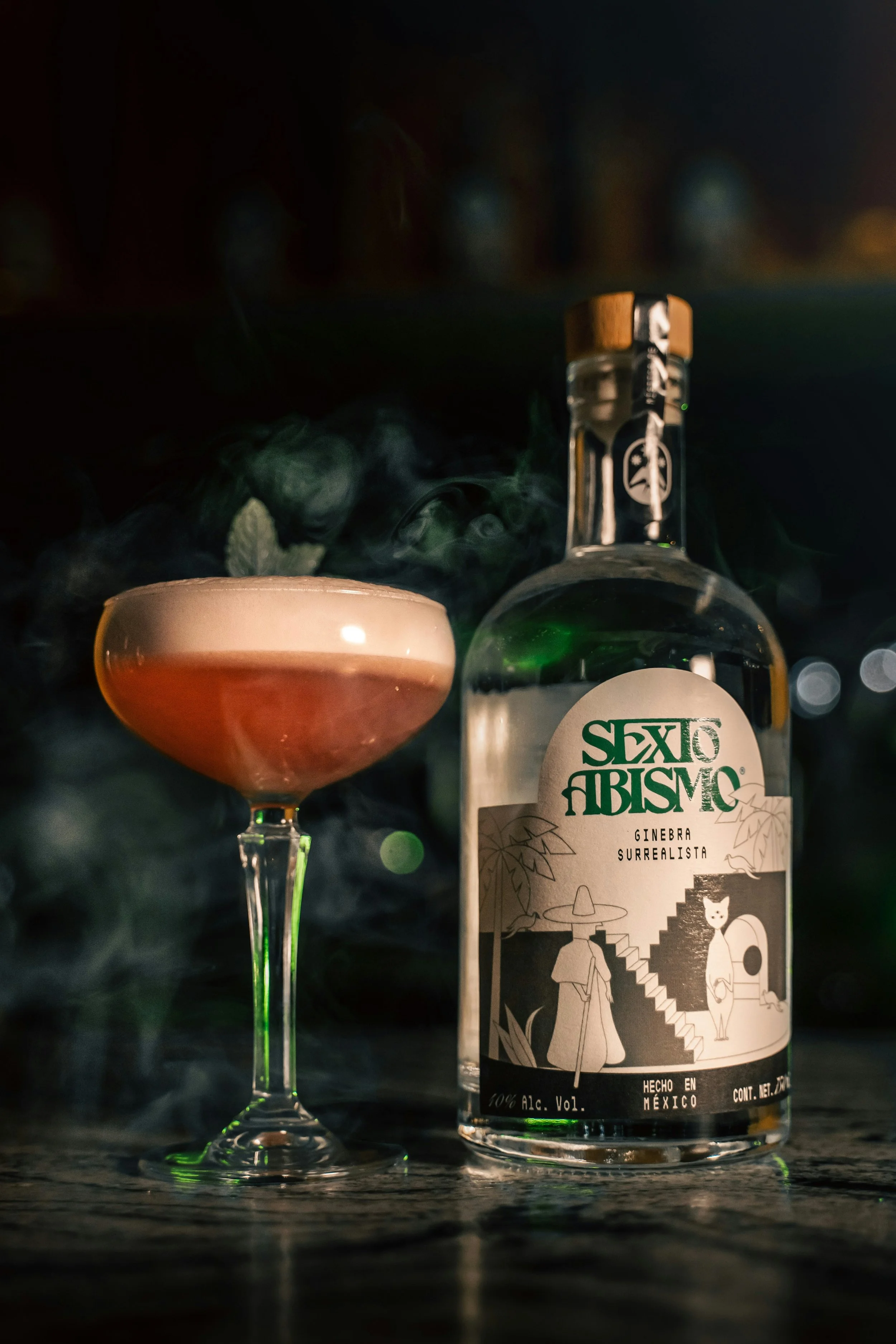 A bottle of Mexican spirit and a glass filled with a pink cocktail with a foamy top, with smoke or vapor around them, on a dark surface.