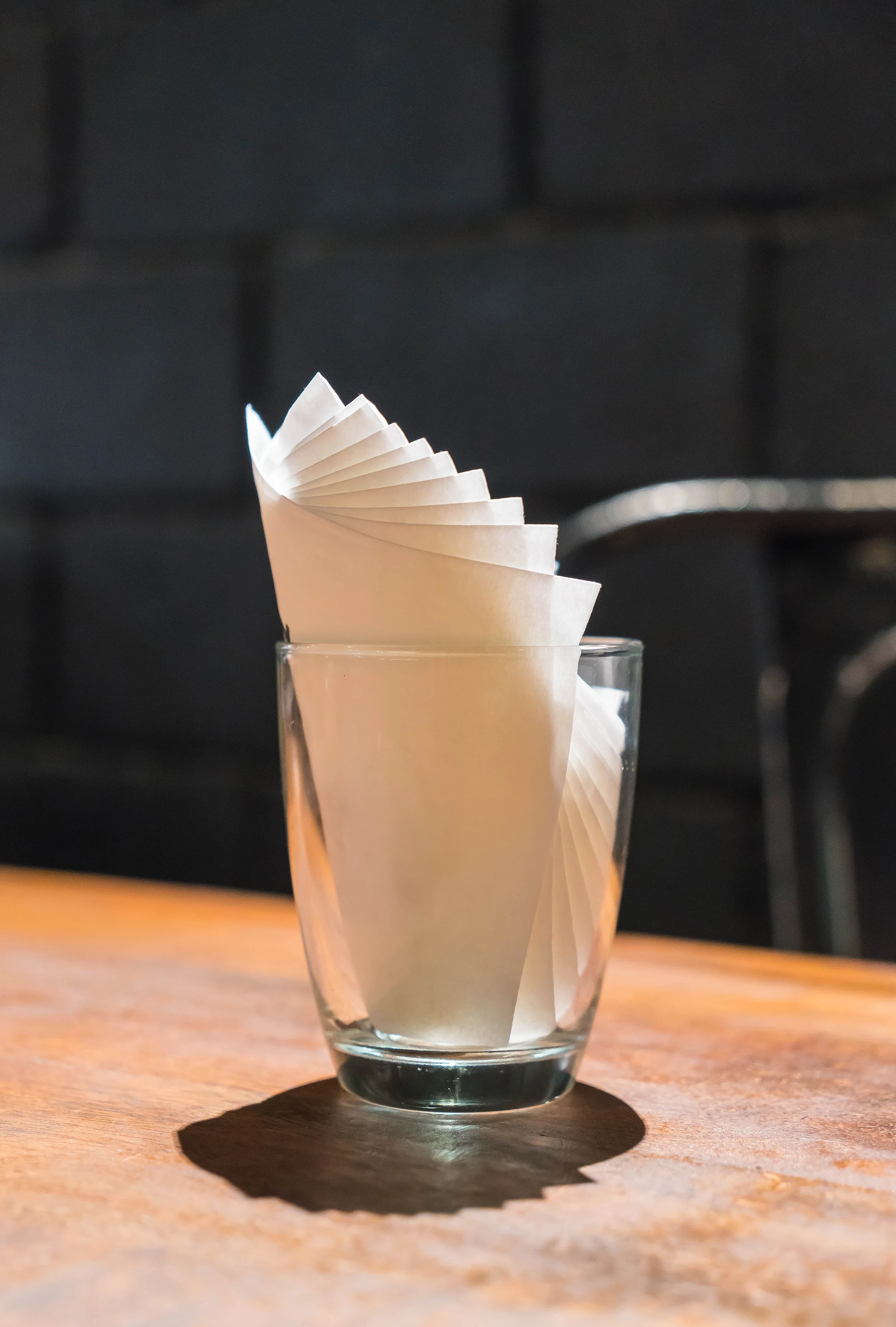 A clear glass filled with folded white paper napkins on a wooden table with a dark background.
