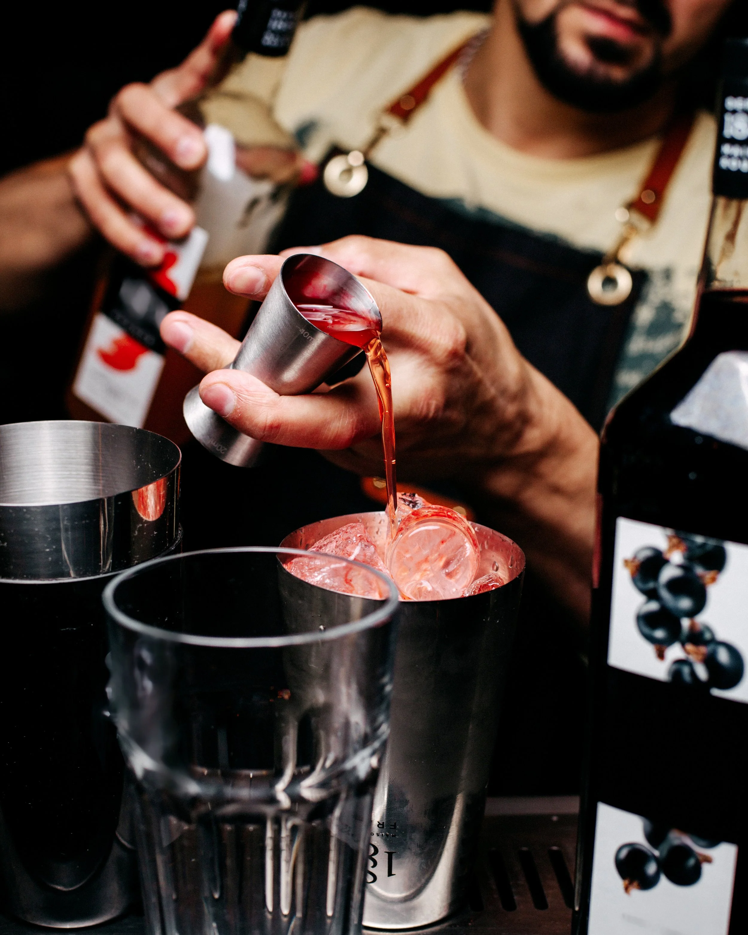 Bartender pouring a red cocktail from a shaker into a glass filled with ice and ice cubes, with cocktail ingredients and glasses on the bar counter.