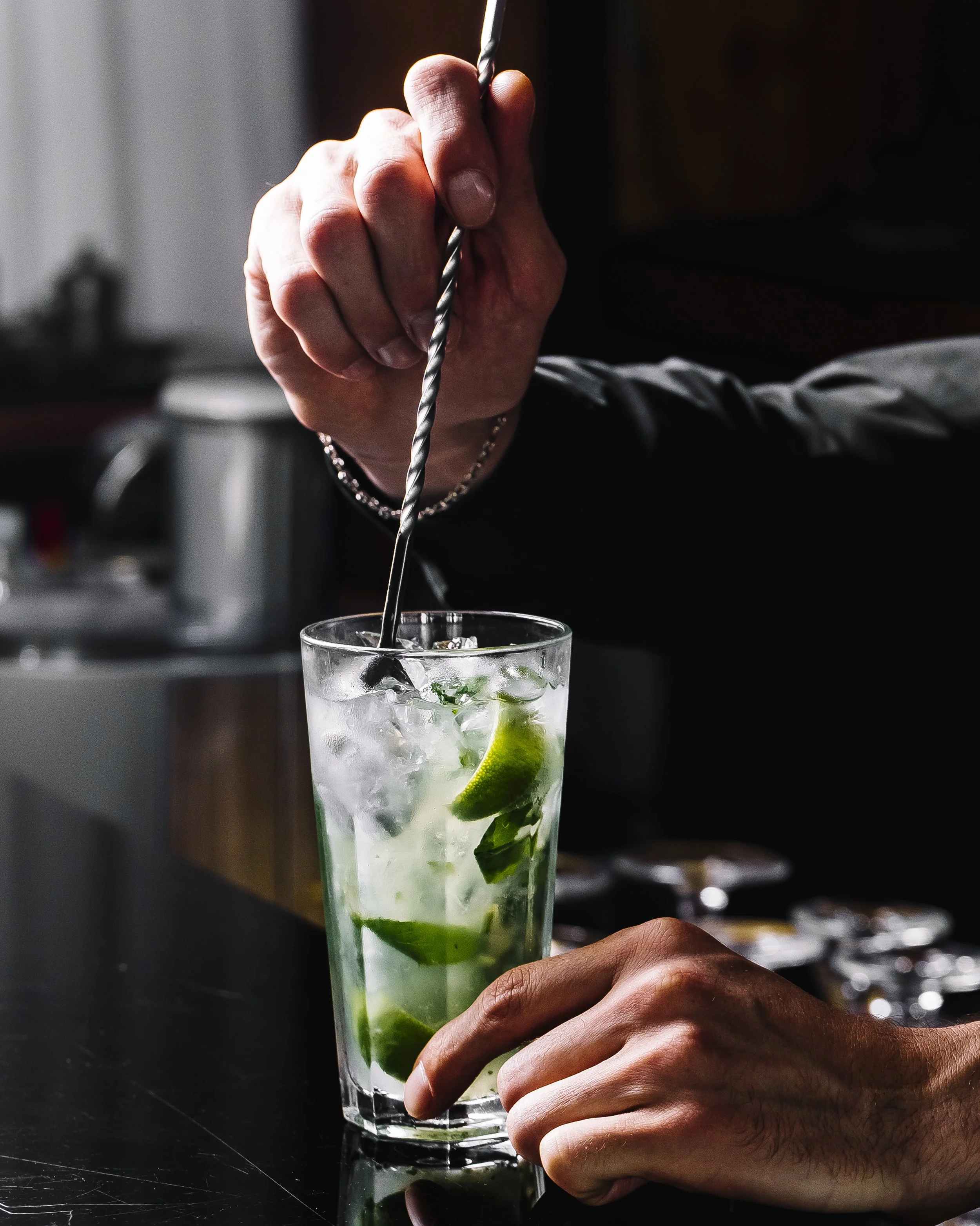 Hand stirring a mojito cocktail with lime and mint in a highball glass on a bar counter.