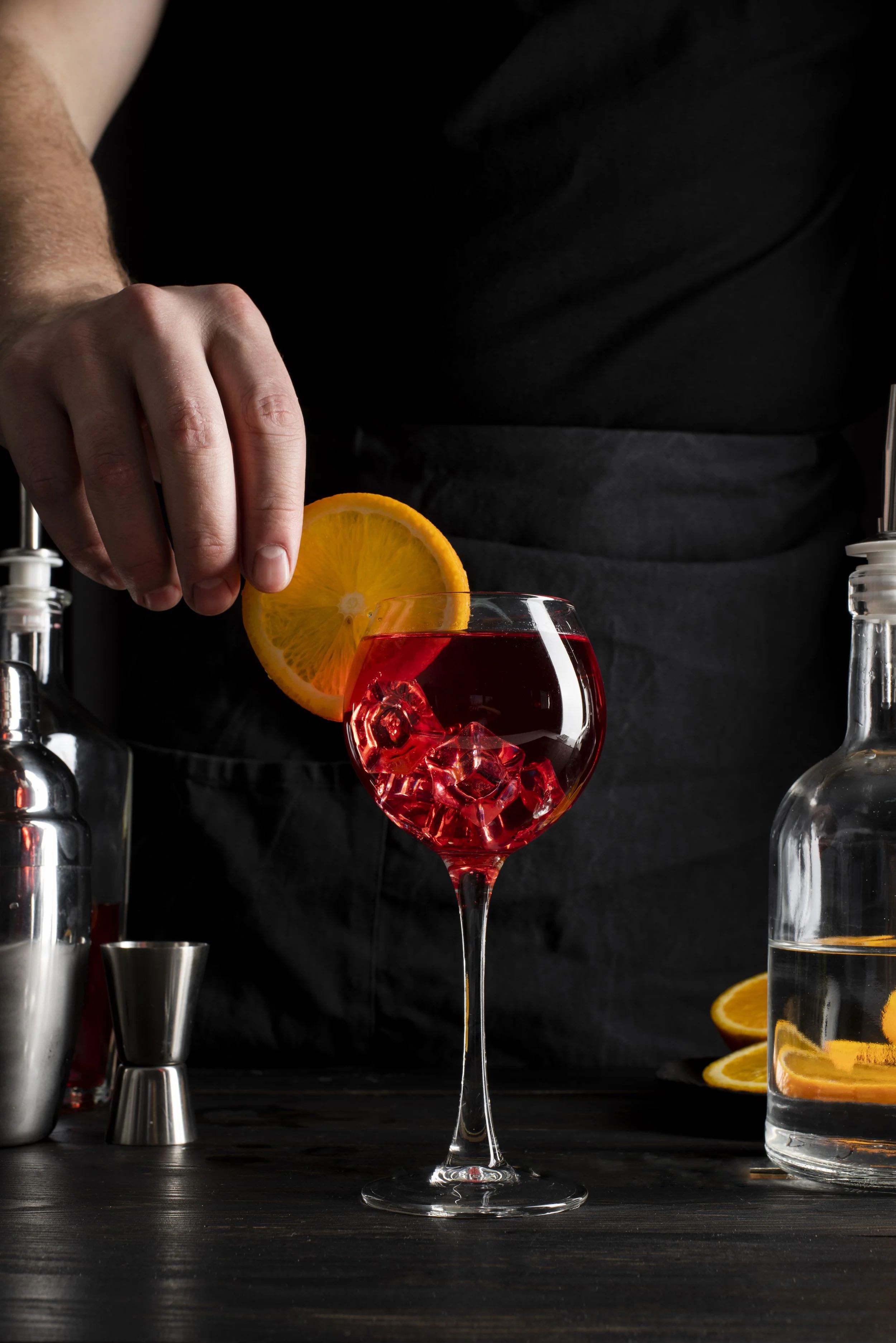 A bartender garnishing a red cocktail with an orange slice in a wine glass filled with ice, surrounded by bar tools and orange slices, against a dark background.