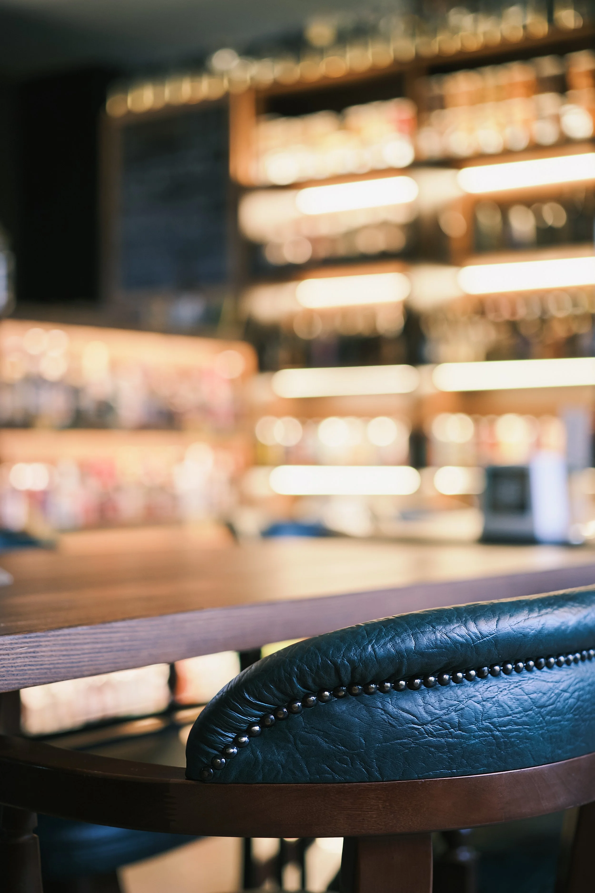 Close-up of a black leather chair with a wooden table in a cozy cafe or restaurant. Blurred shelves with bottles and a chalkboard menu in the background.