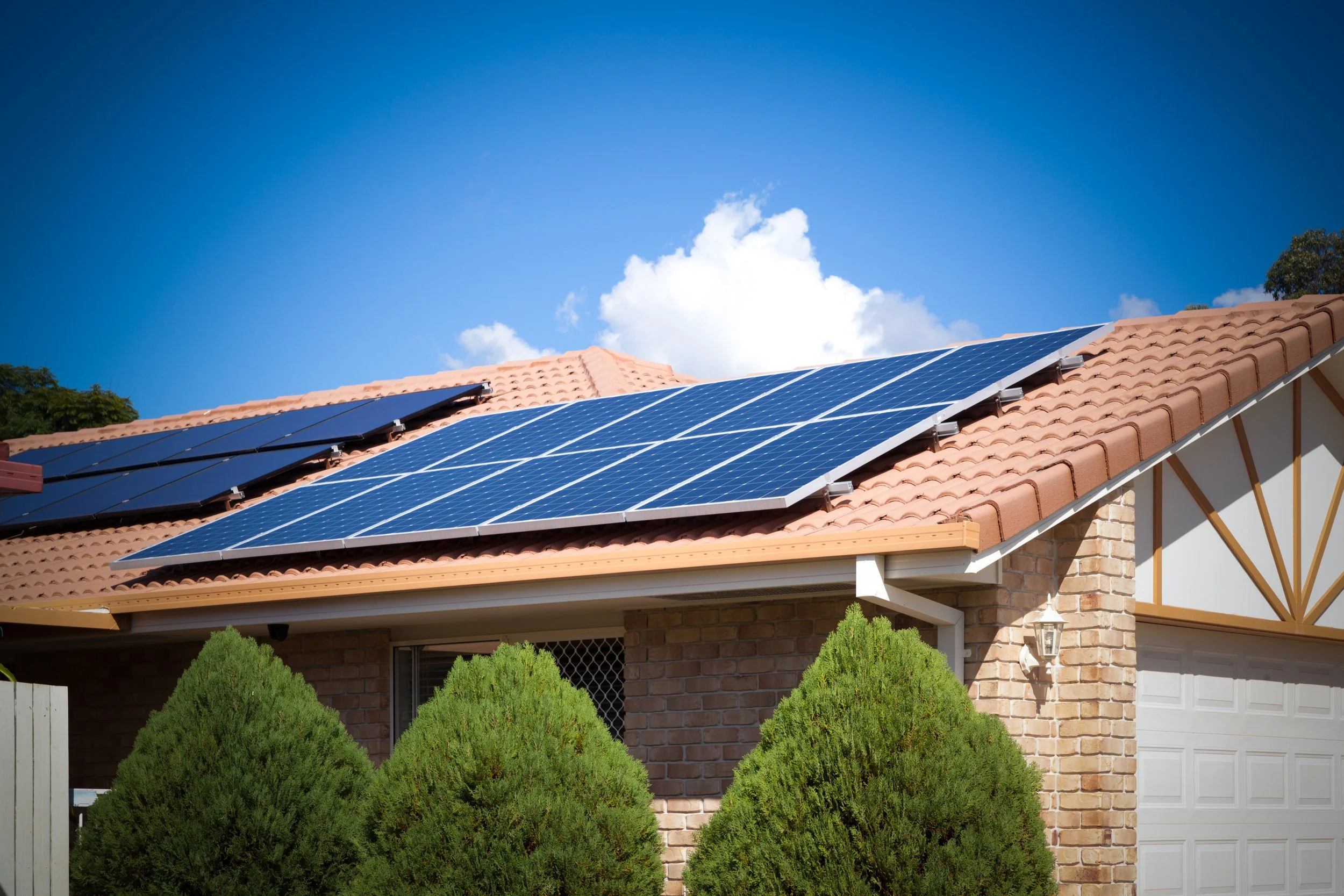 Solar panels installed on a brick house roof with bushes in the front and a blue sky with clouds in the background.