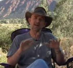 A man wearing a wide-brimmed hat and gray shirt, sitting outdoors with a mountain and trees in the background, speaking or gesturing with his hands.