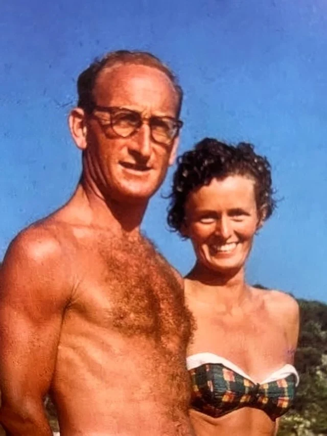 A shirtless man with glasses and a woman in a striped bikini top standing outdoors under a clear blue sky.